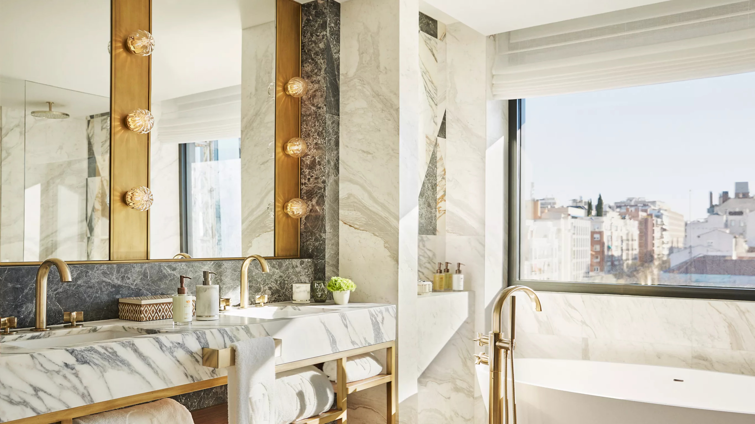 A bathroom vanity with gold mirror frame, marble countertop, and windows with city views