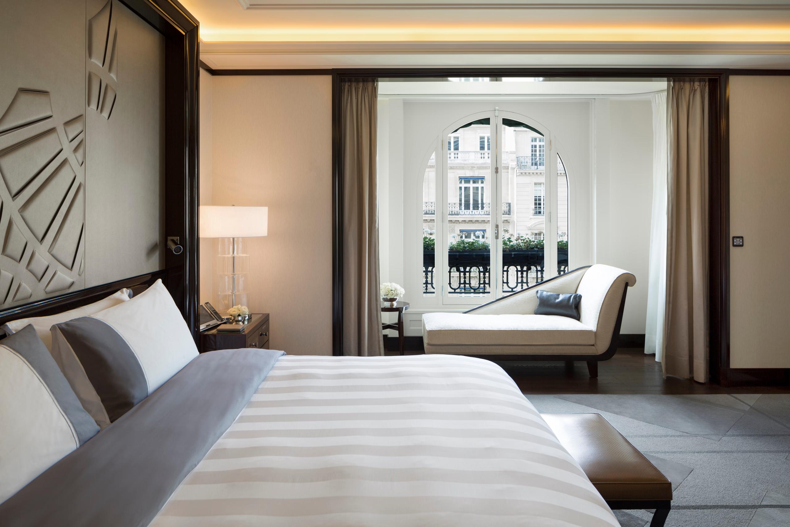 Hotel suite with bed facing a window, warm lighting, wall sconces, and period furnishings