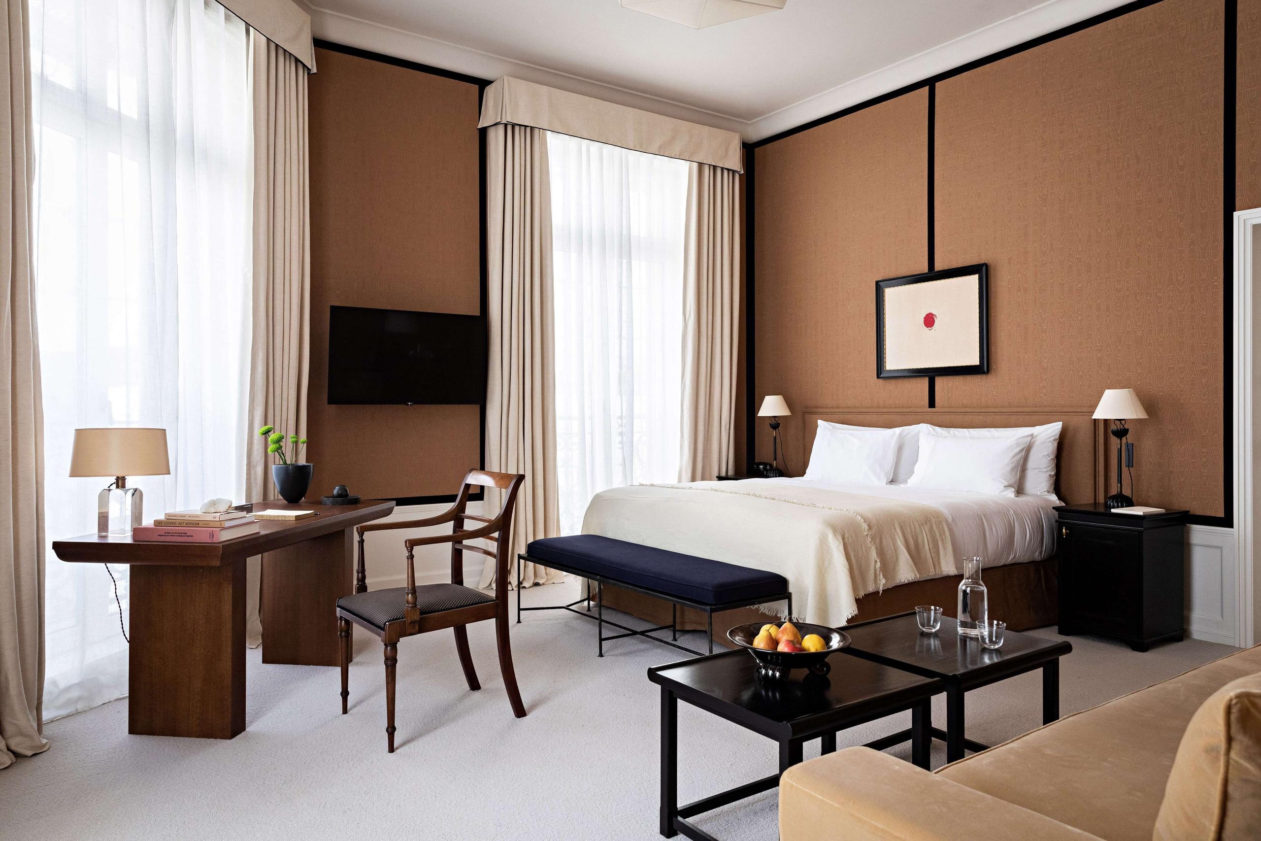 A bedroom with tan and brown tones, wood furnishings, and tall windows