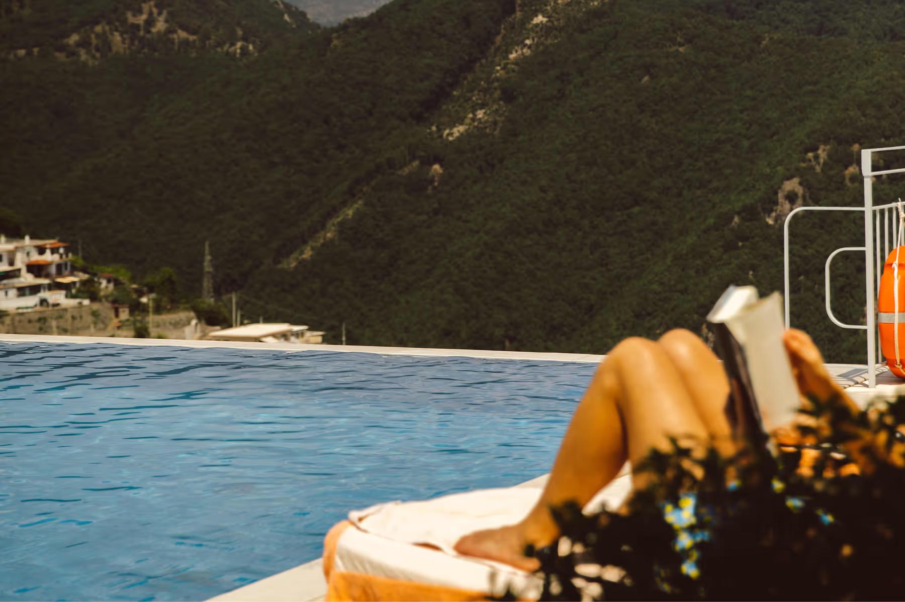 Caruso: blue infinity pool edge with yellow lounger overlooking rocky coastline and terraced village below