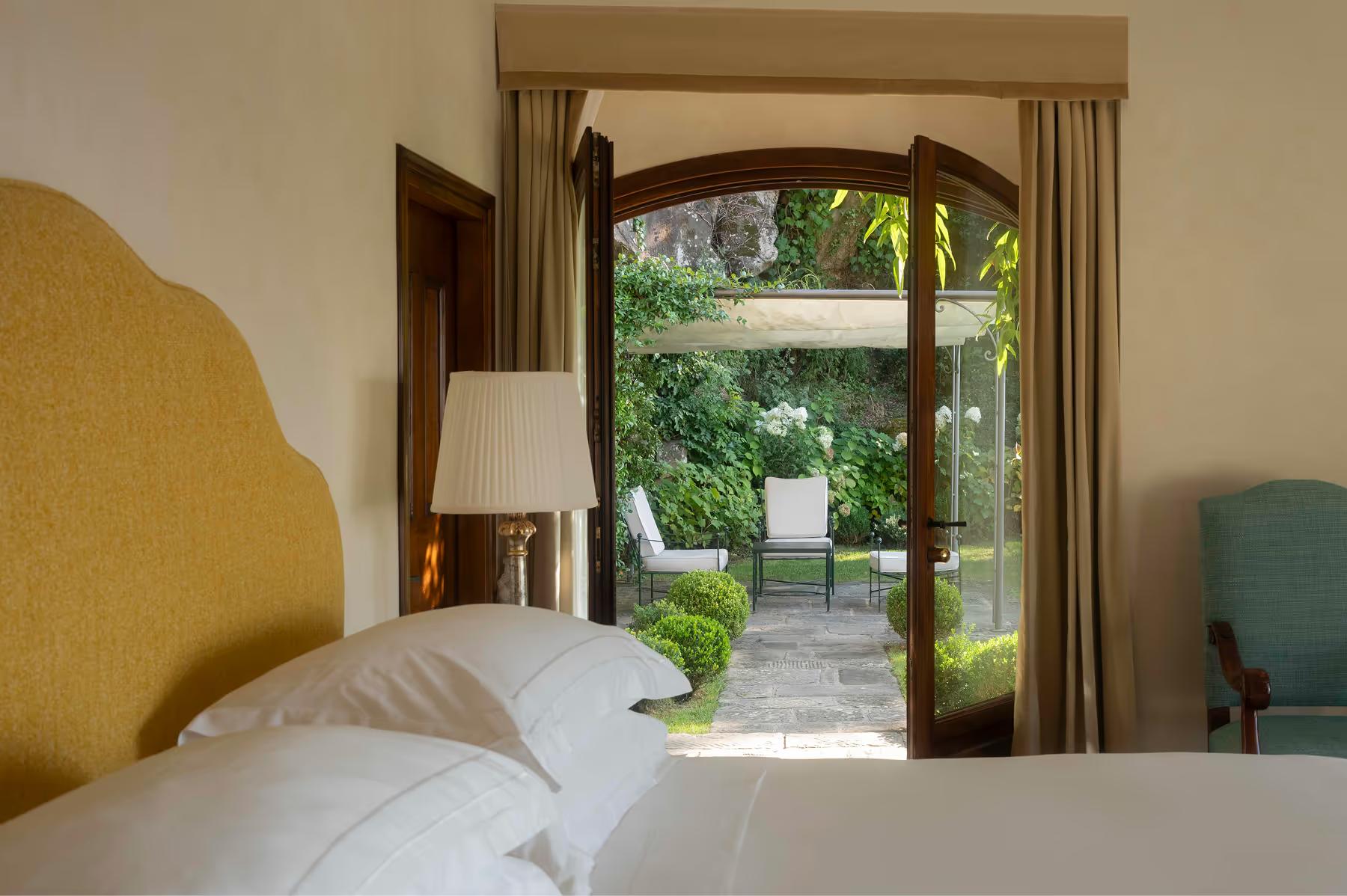 Villa San Michele: a bright bedroom with arched doorway opening to a garden view, mustard accent chair, and terracotta floor