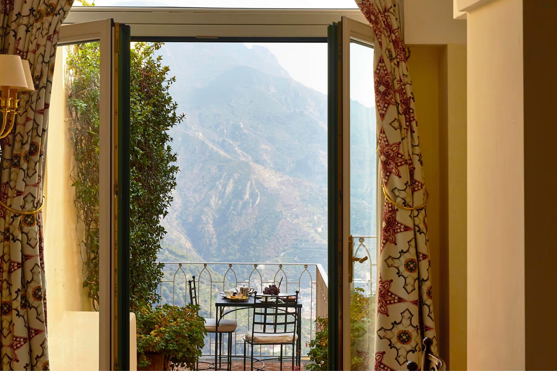 Caruso: room window framing coastal mountains with floral patterned curtains and wrought-iron balcony railing