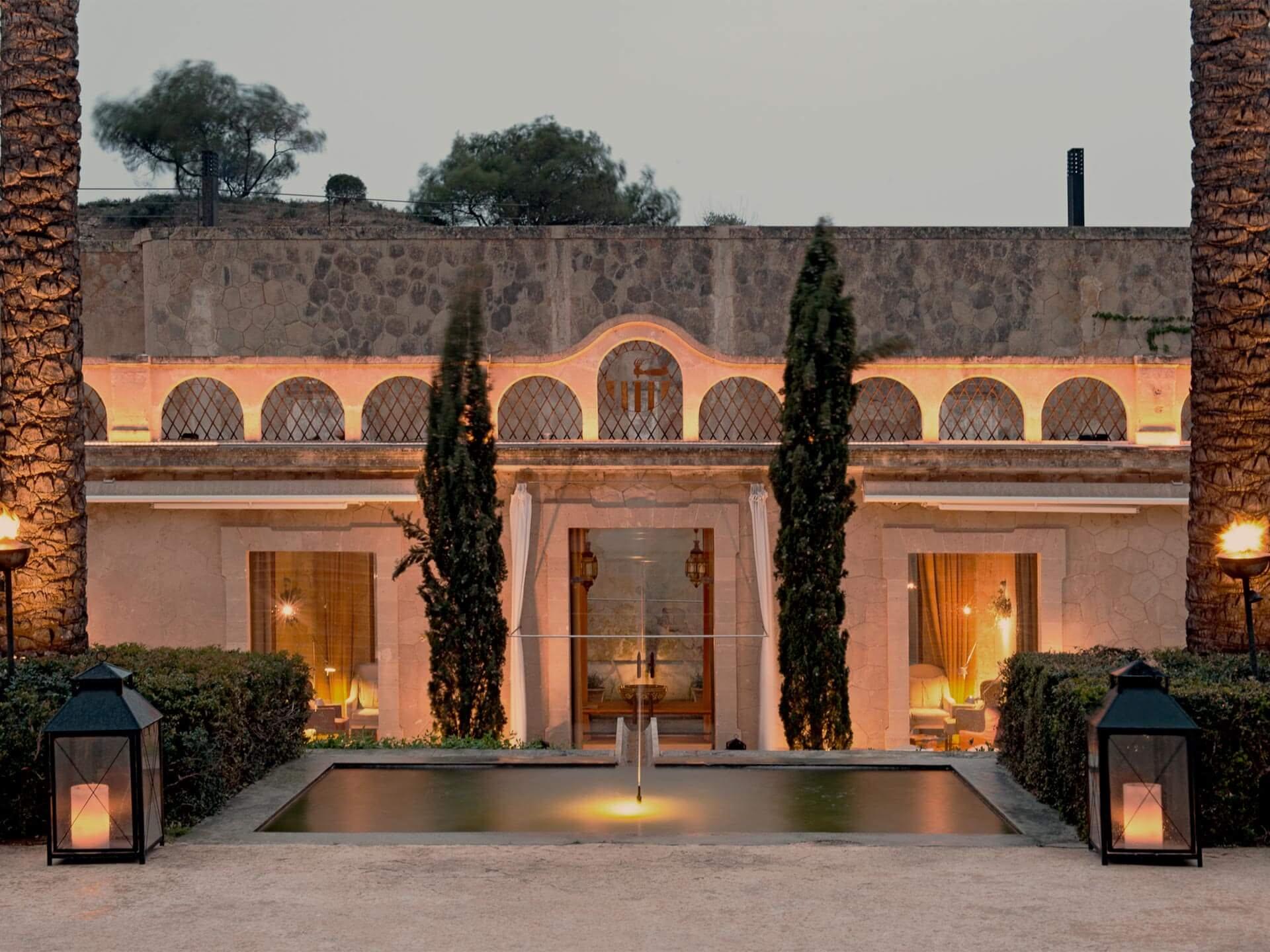 A cream and terracotta fortress facade with rounded arched windows, flanked by tall cypress trees and lanterns