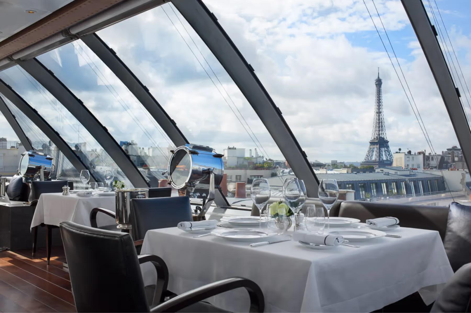 Rooftop dining terrace with glass panels, white tablecloths, and panoramic views of the Eiffel Tower