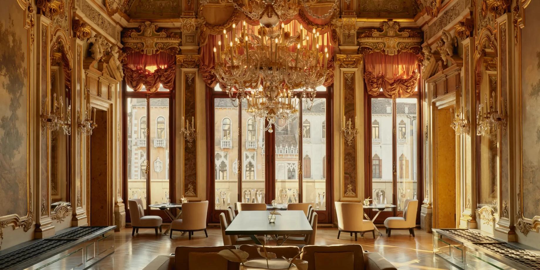 A grand gilded Venetian salon with a large crystal chandelier and tall arched windows over the Grand Canal