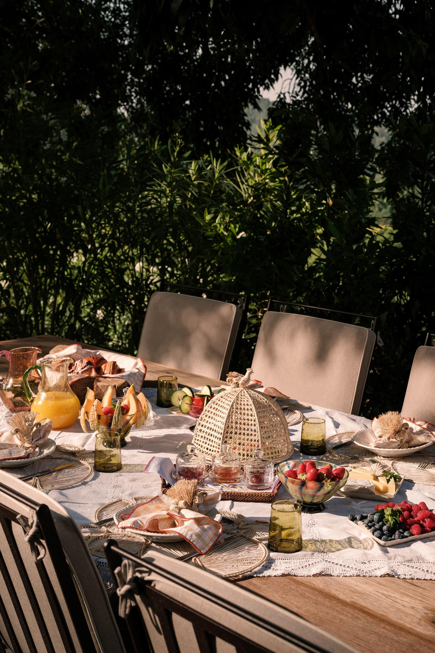 La Bergerie: outdoor breakfast table set with pastries and fruit beneath woven shade sail, lush green foliage, dappled light