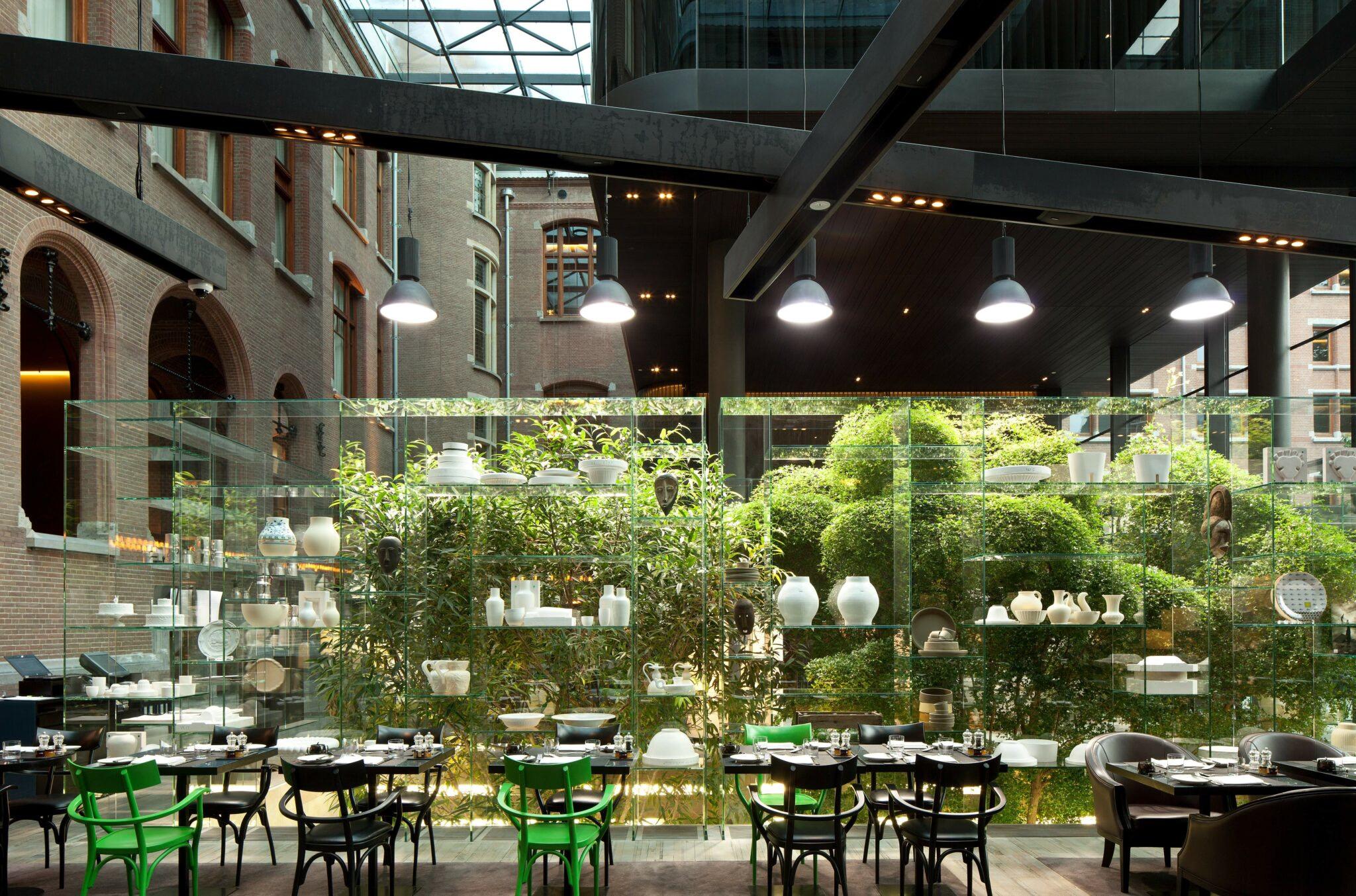Mandarin Oriental Conservatorium, Amsterdam: restaurant tables and green chairs in the glass-roofed atrium, with brick facades and glass shelves of ceramics and plants