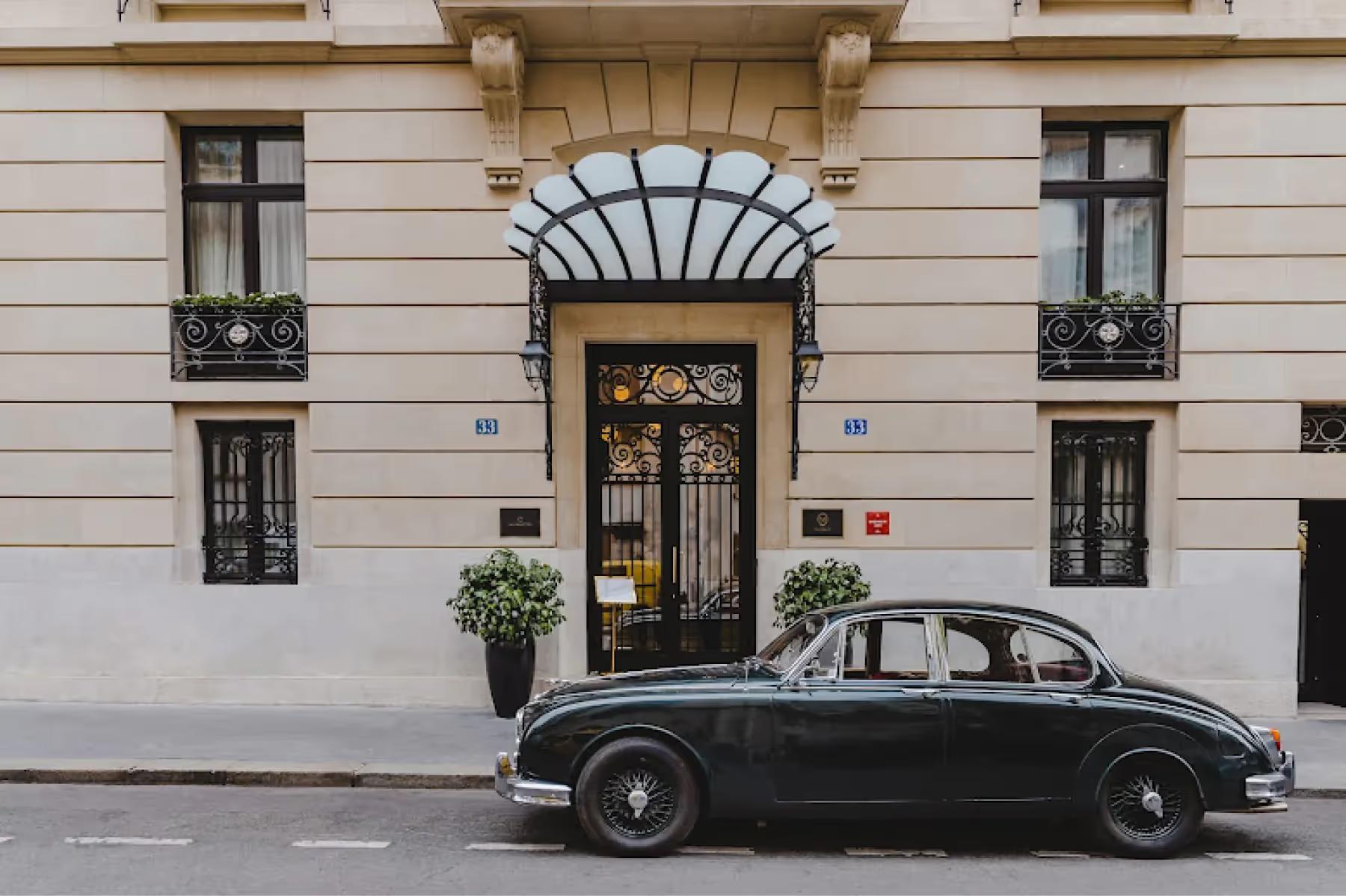 A classical beige facade with arched entry, black-trimmed windows, and a black vintage car parked outside