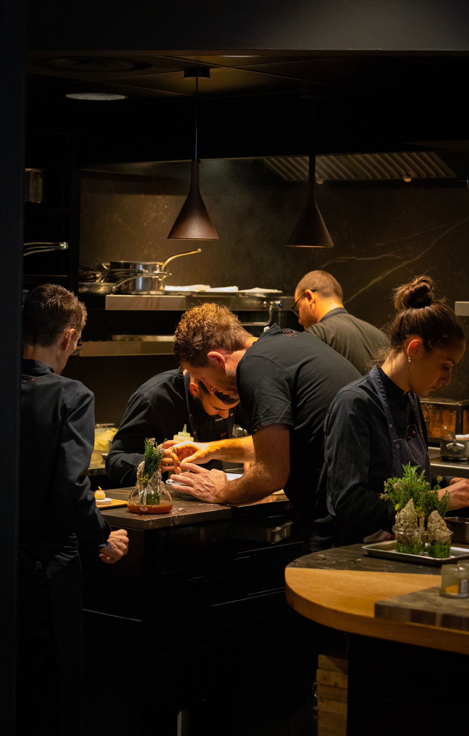 Chefs plating dishes together at the open kitchen counter of AM par Alexandre Mazzia, under pendant lights