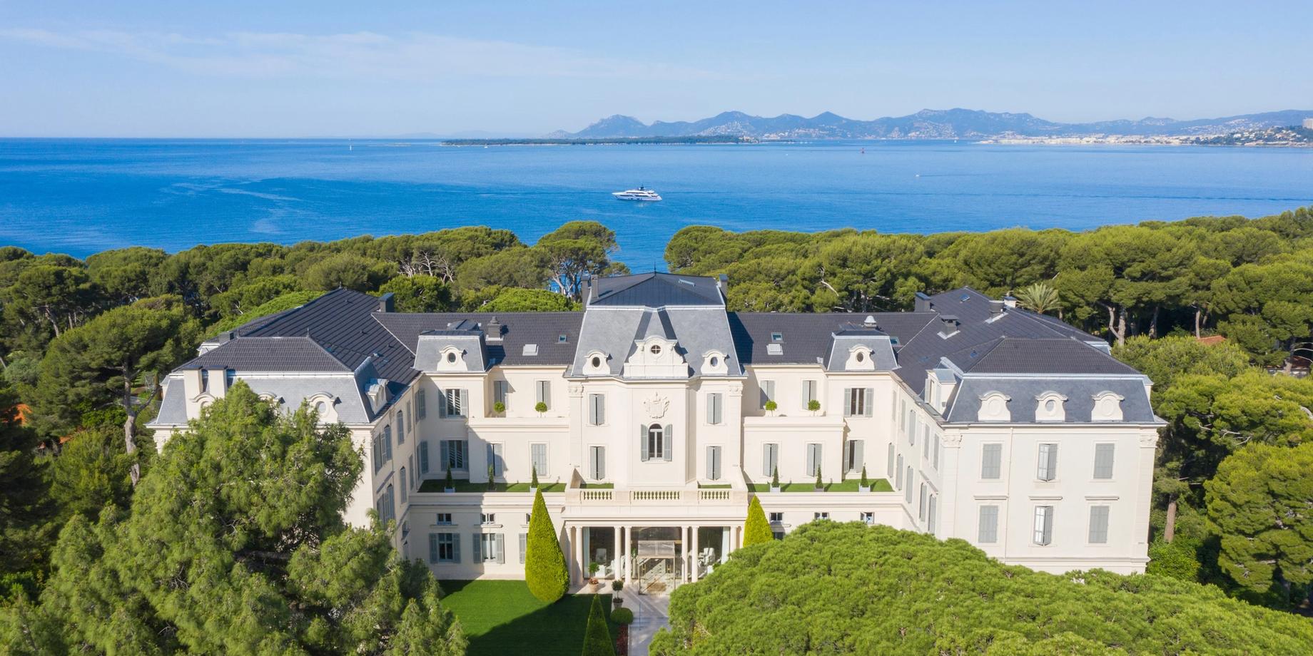 A sweeping aerial view of a whitewashed stone mansion with manicured grounds, pine trees, and the Mediterranean coastline in the background