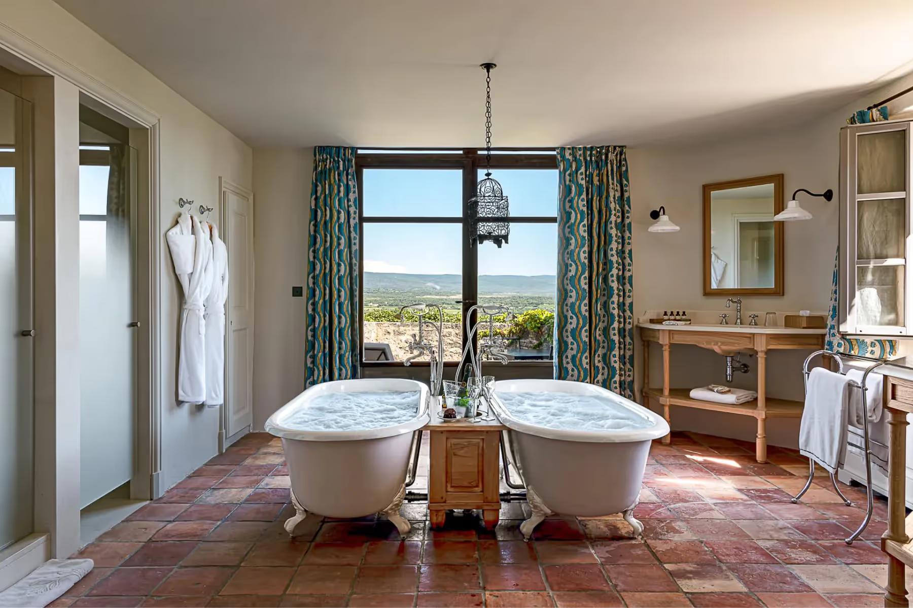 Hôtel Crillon le Brave: a spa-like bathroom with two white bathtubs, terracotta tile floor, and countryside views through large windows