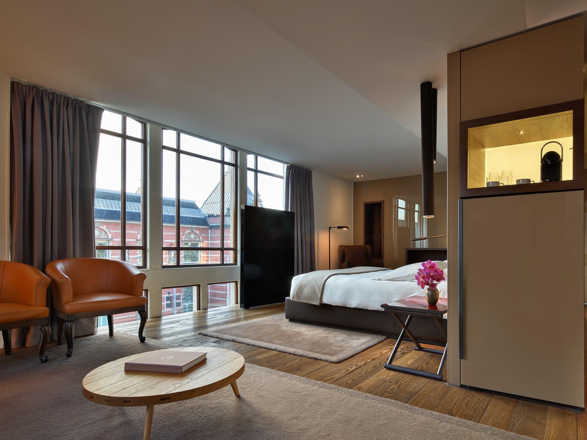 Mandarin Oriental Conservatorium, Amsterdam: bedroom with tall windows facing a red-brick building, two armchairs, and an oval wooden coffee table