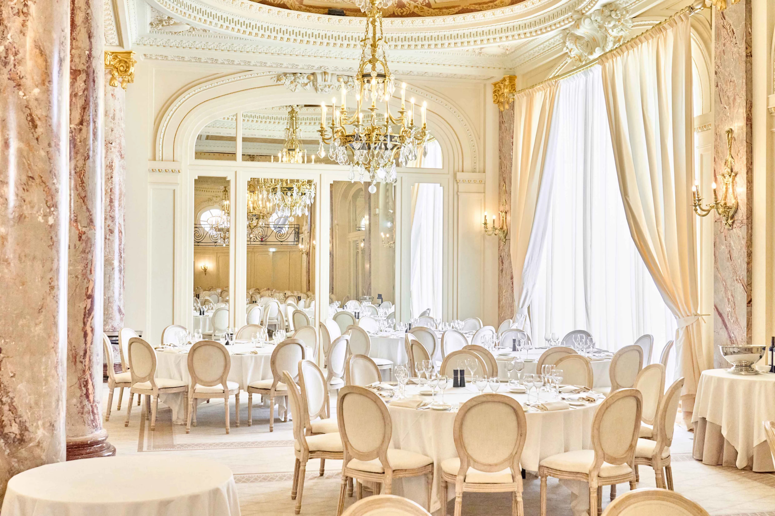 Carlton Cannes: elegant dining room with gilt trim, cream-colored walls, white tablecloths, and pale wood chairs under a crystal chandelier