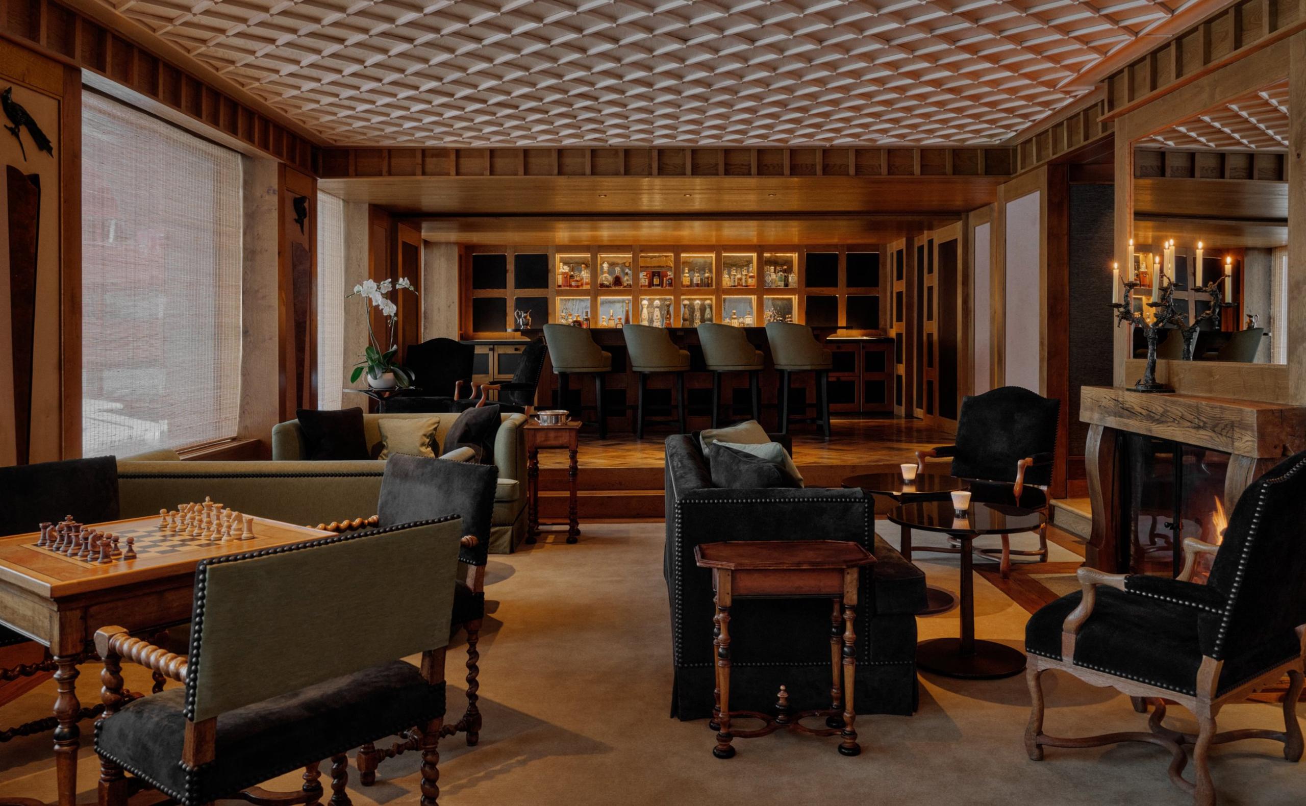 Aman Le Mélézin: games room with a fireplace, chess table, leather chairs, and a lit bar beneath a coffered ceiling