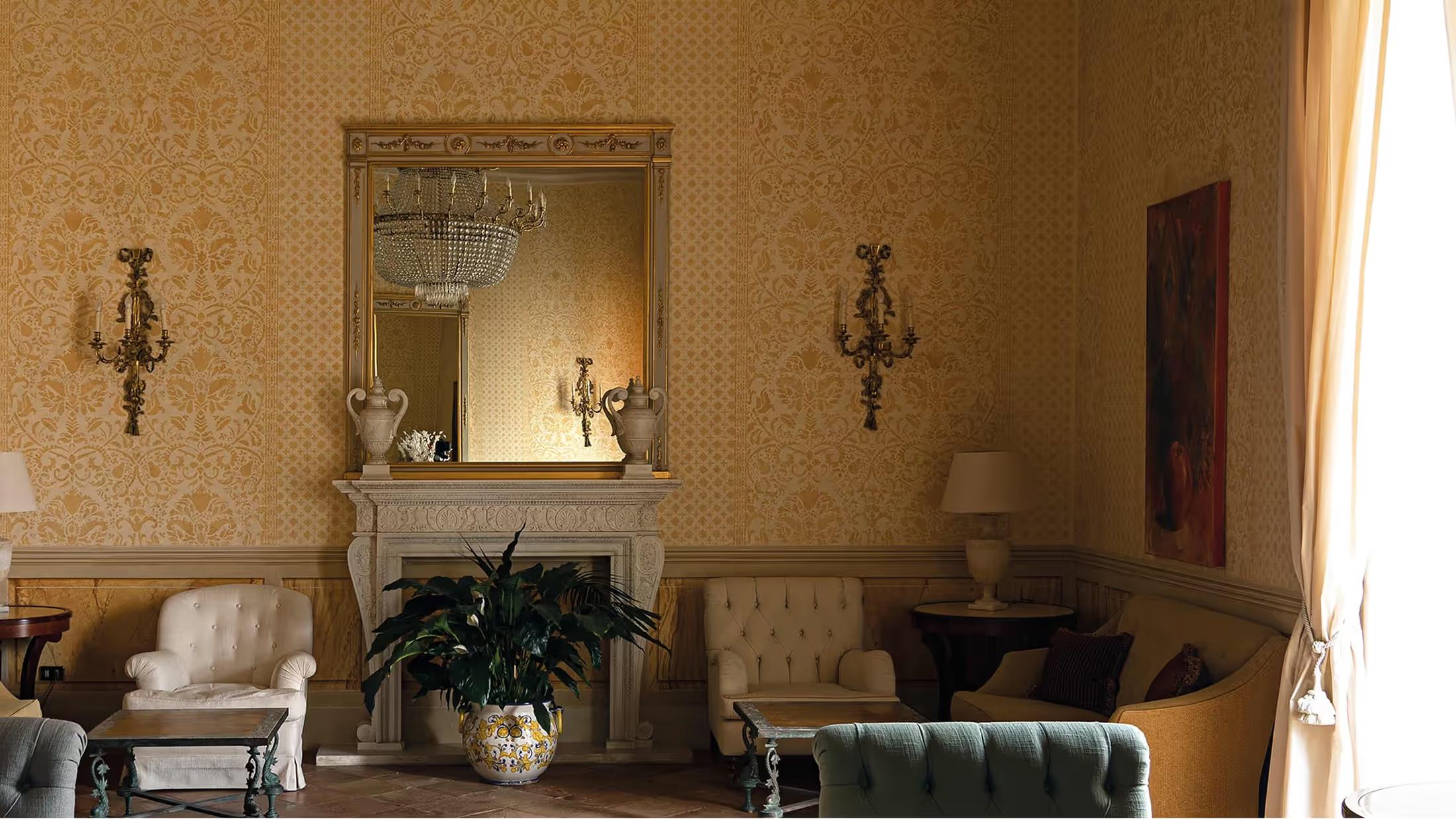A graceful room with golden ochre walls, a fireplace with marble mantel, mirror, and plant arrangements beneath wall sconces