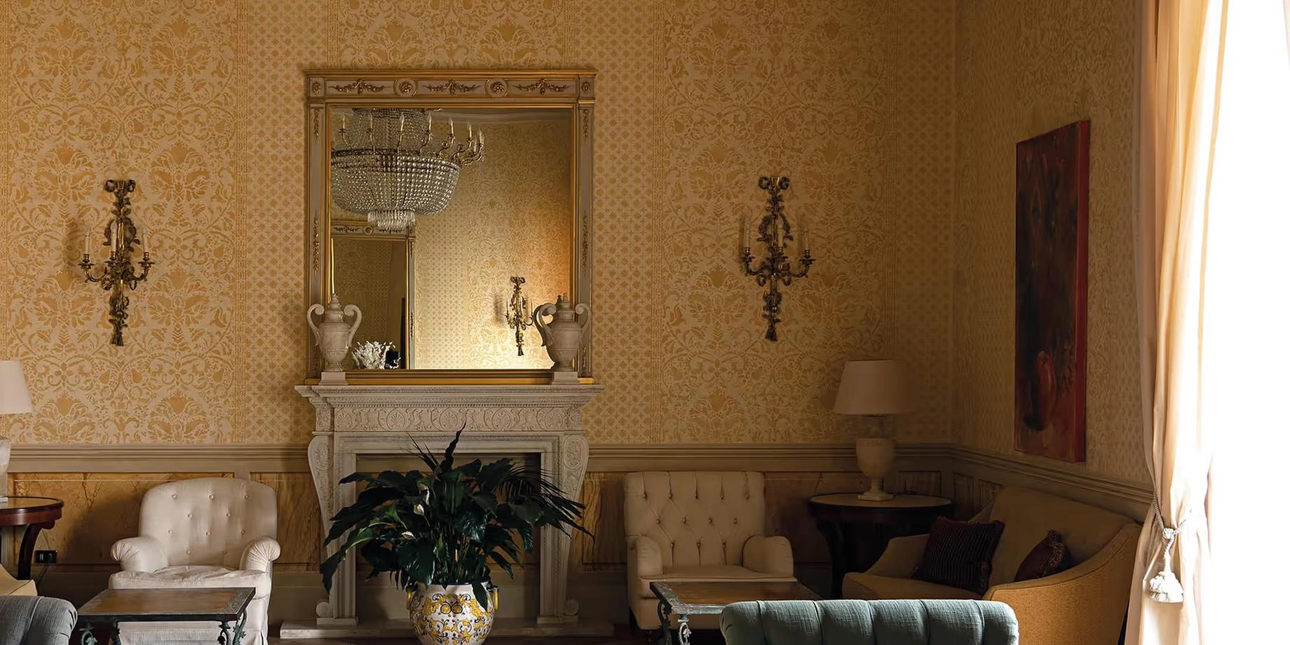 A graceful room with golden ochre walls, a fireplace with marble mantel, mirror, and plant arrangements beneath wall sconces
