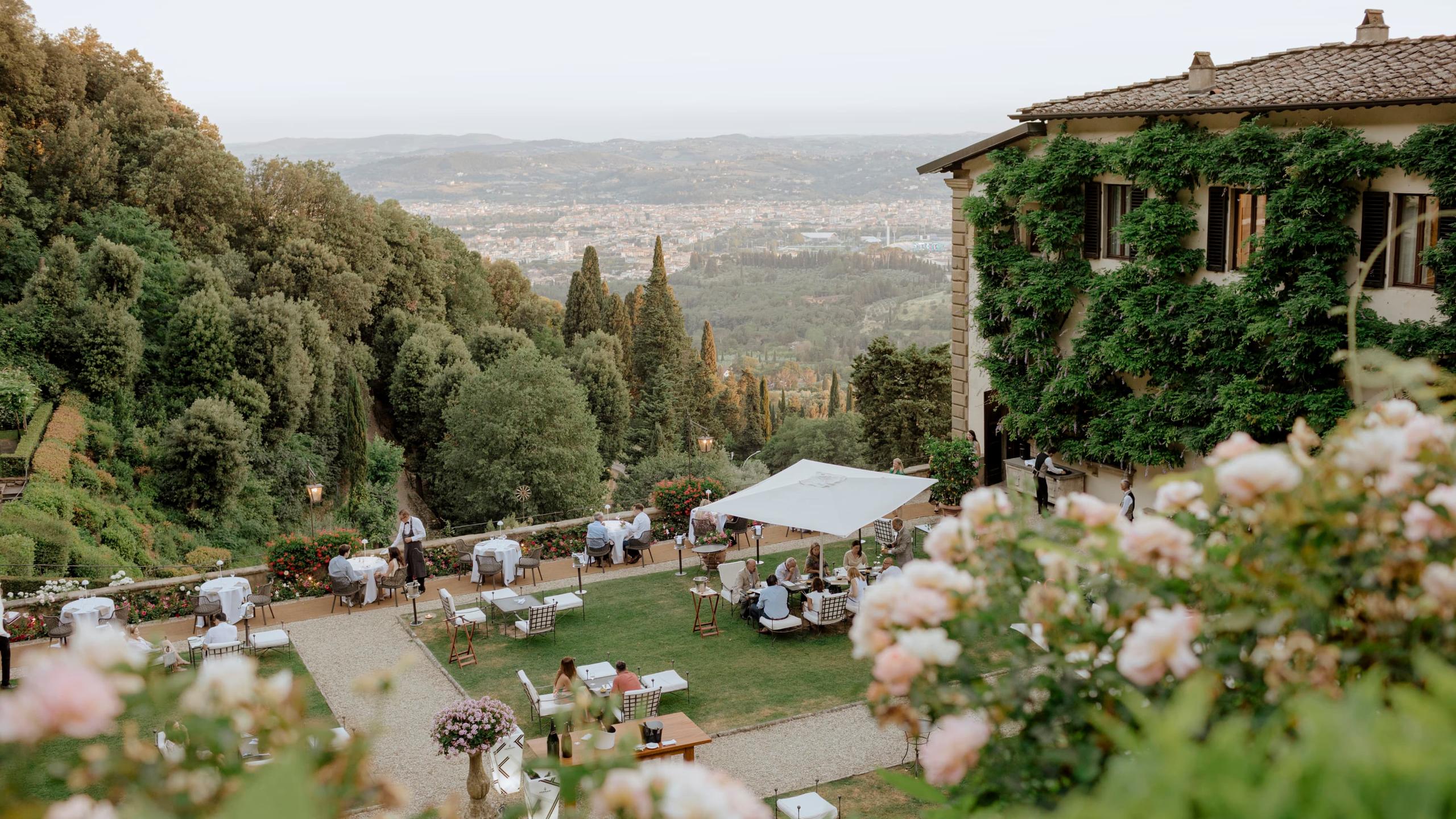 Villa San Michele: an afternoon garden party with guests at scattered tables, white umbrellas, and valley views beyond