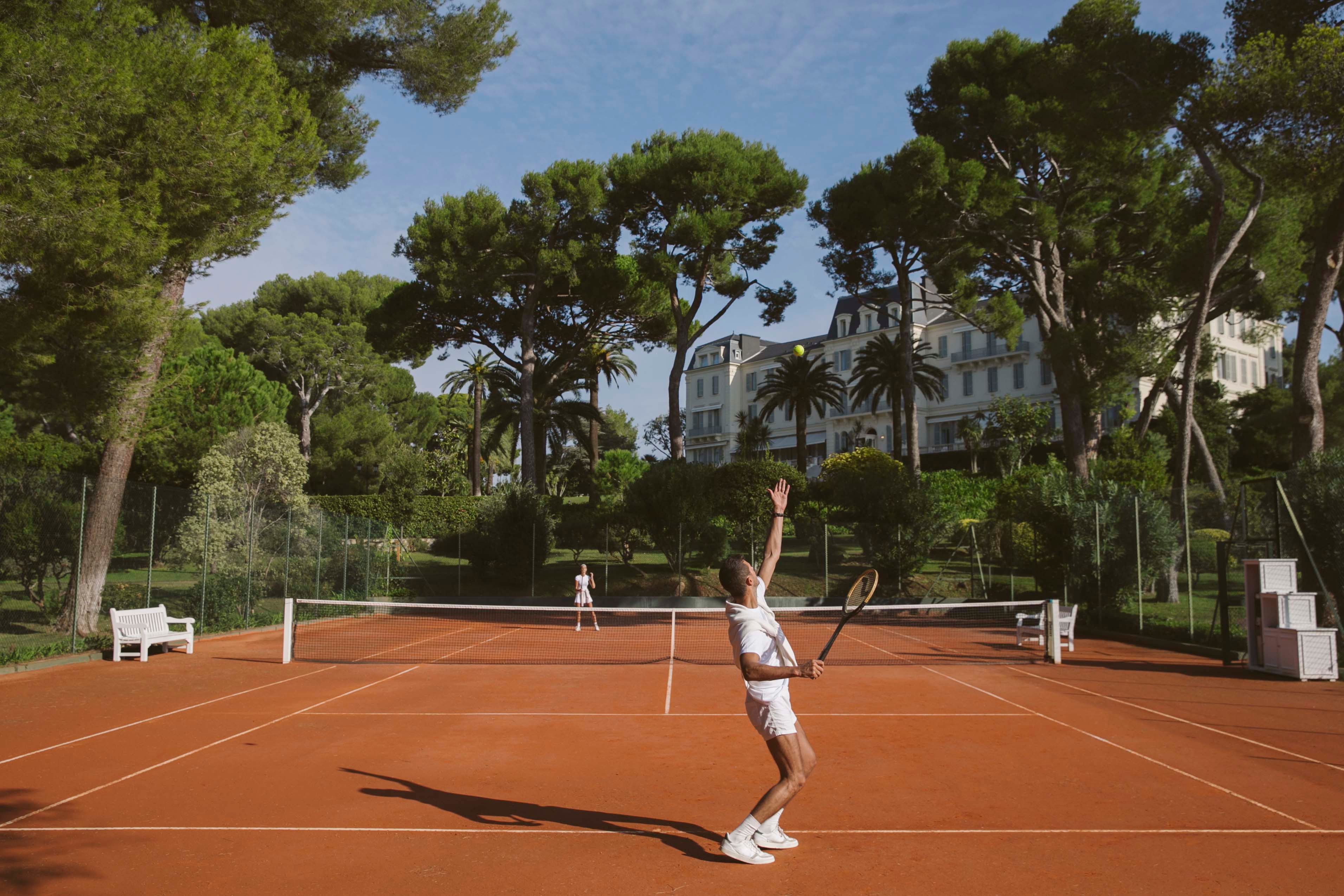 A player serving on a clay court under the pines at the Hôtel du Cap-Eden-Roc