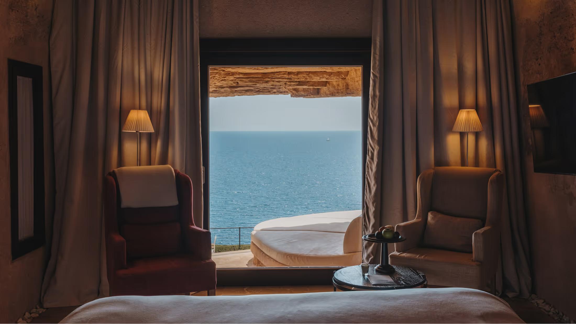 A darkened room frames a calm turquoise sea, fringed in bronze wooden trim and soft cushioned furniture