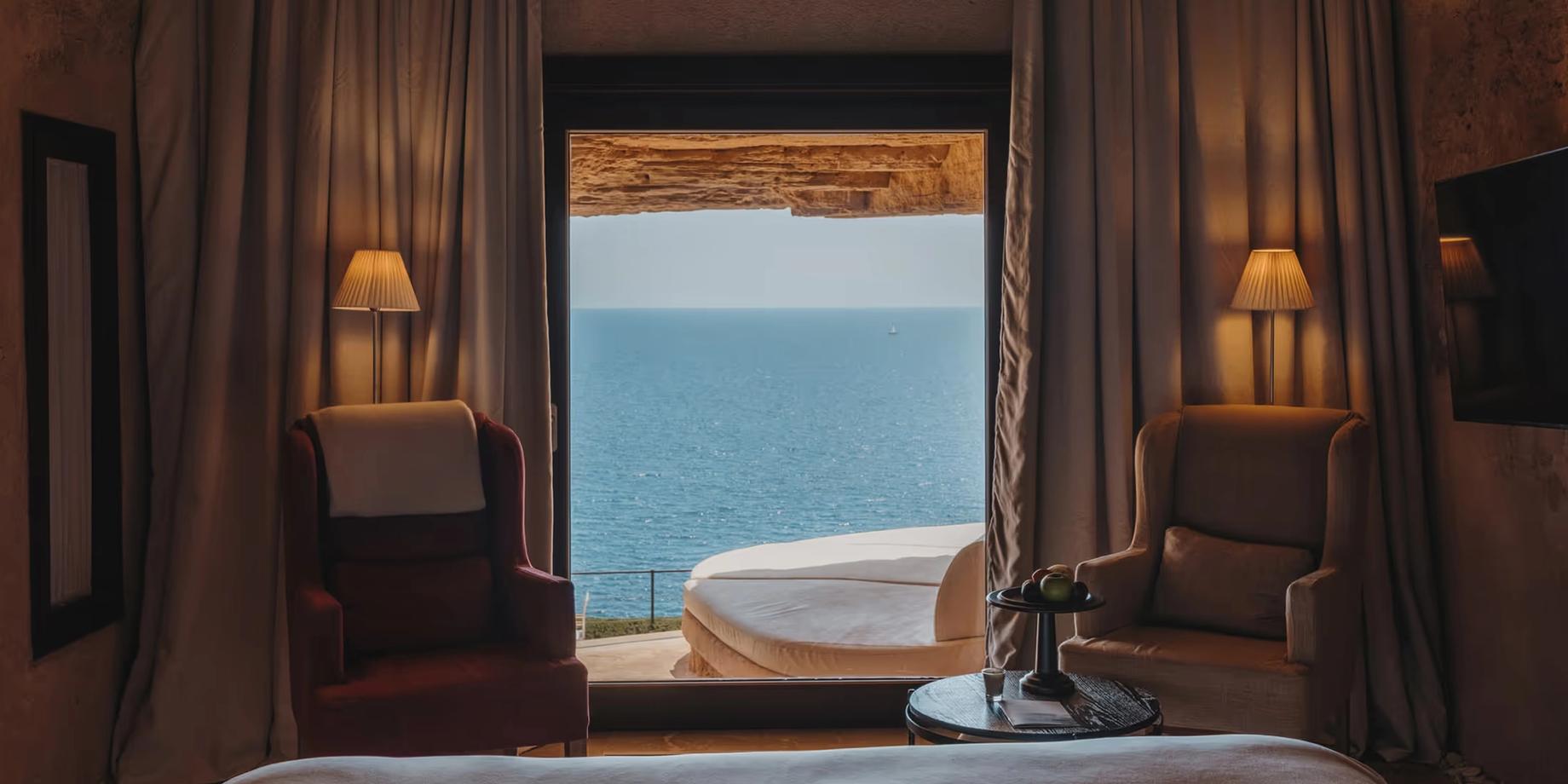 A darkened room frames a calm turquoise sea, fringed in bronze wooden trim and soft cushioned furniture