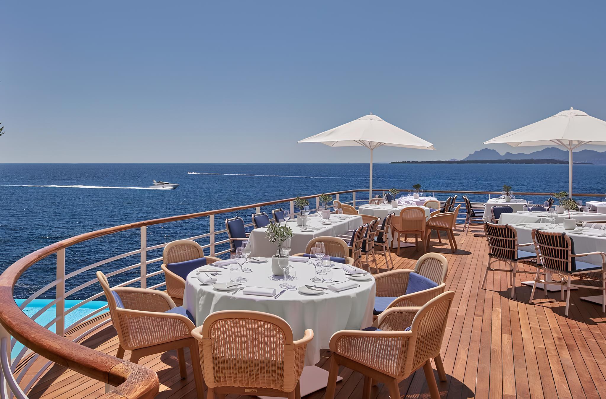 A terrace restaurant with round tables, wooden chairs, white umbrellas, and sea views