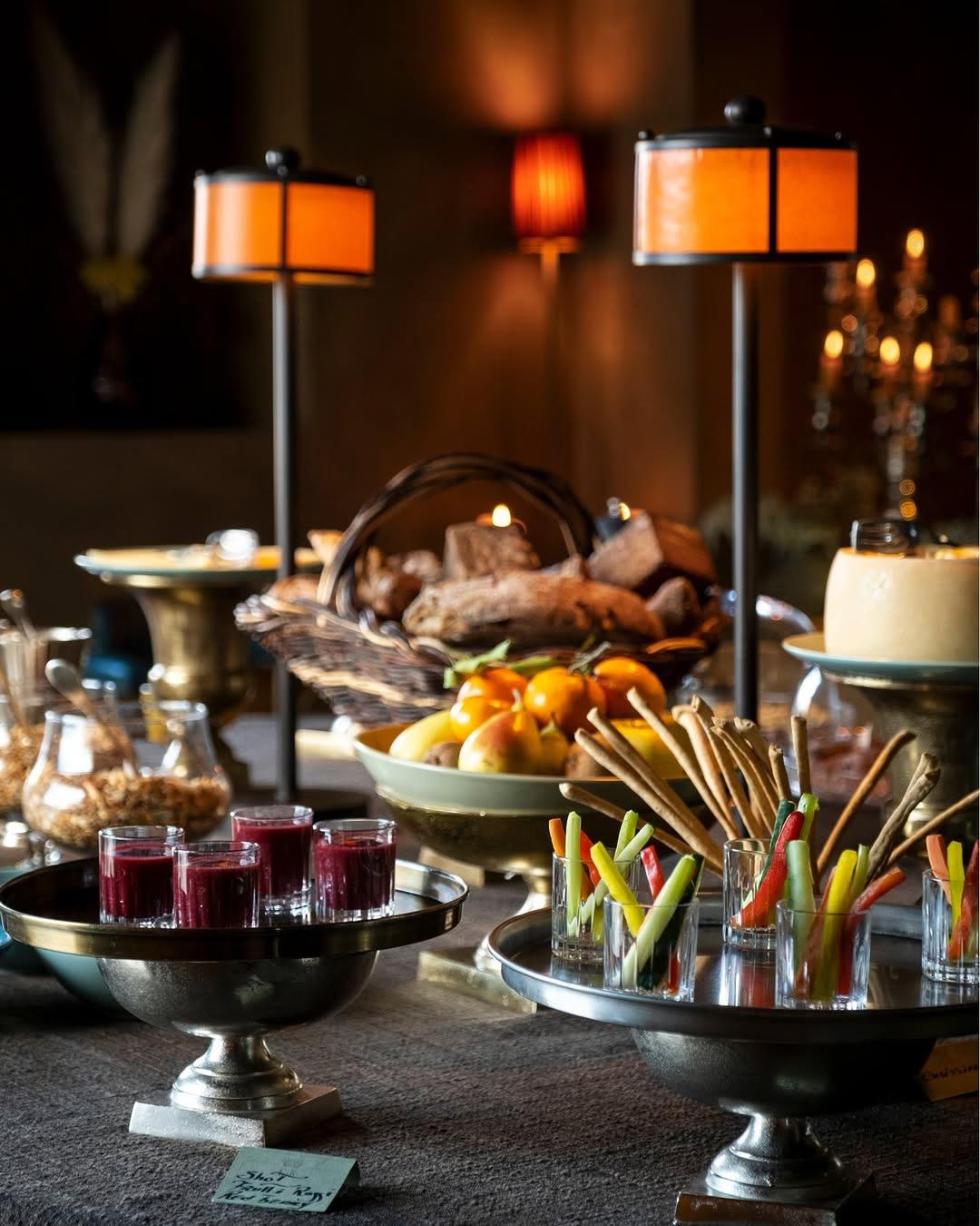 A silver tiered table spread of fruit, bread, and crudites lit by candlelight and warm lamps