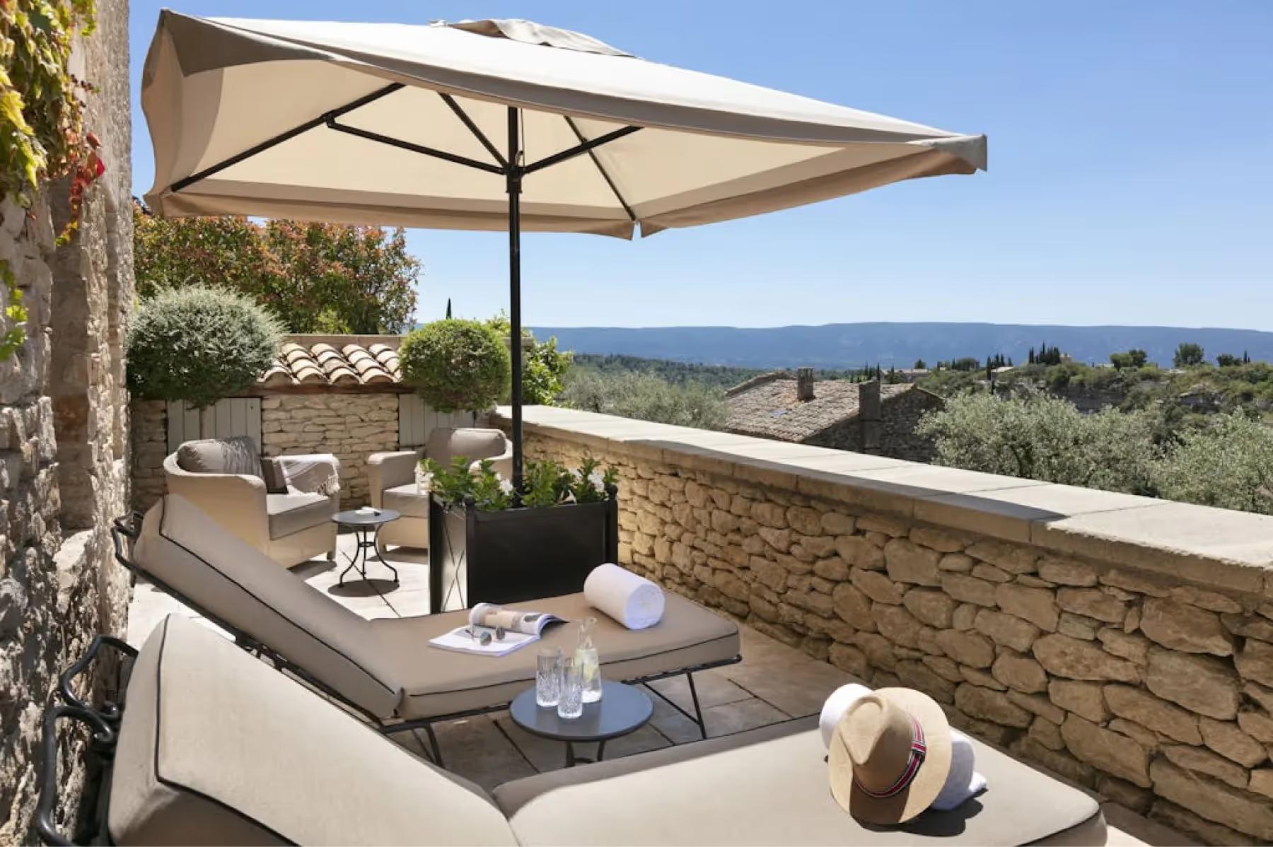 Airelles Gordes, La Bastide: shaded terrace with loungers and stone wall overlooking olive groves and distant mountains
