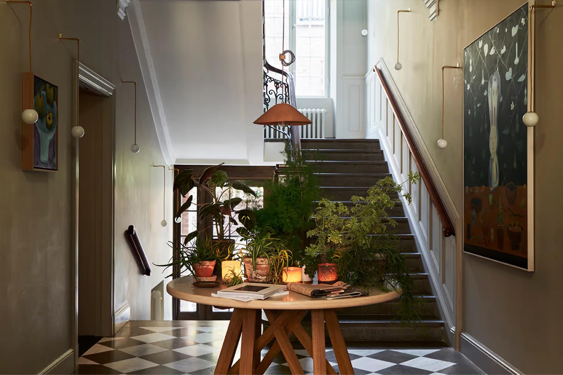 A cream hallway with potted plants on a round wooden table beneath a staircase