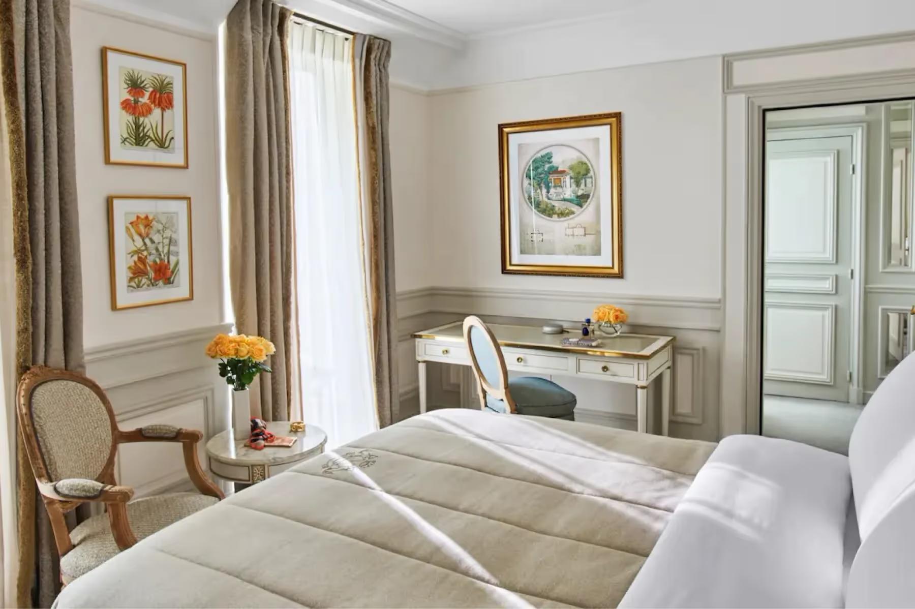 A suite with queen bed, pale furnishings, framed prints, and tall windows with views