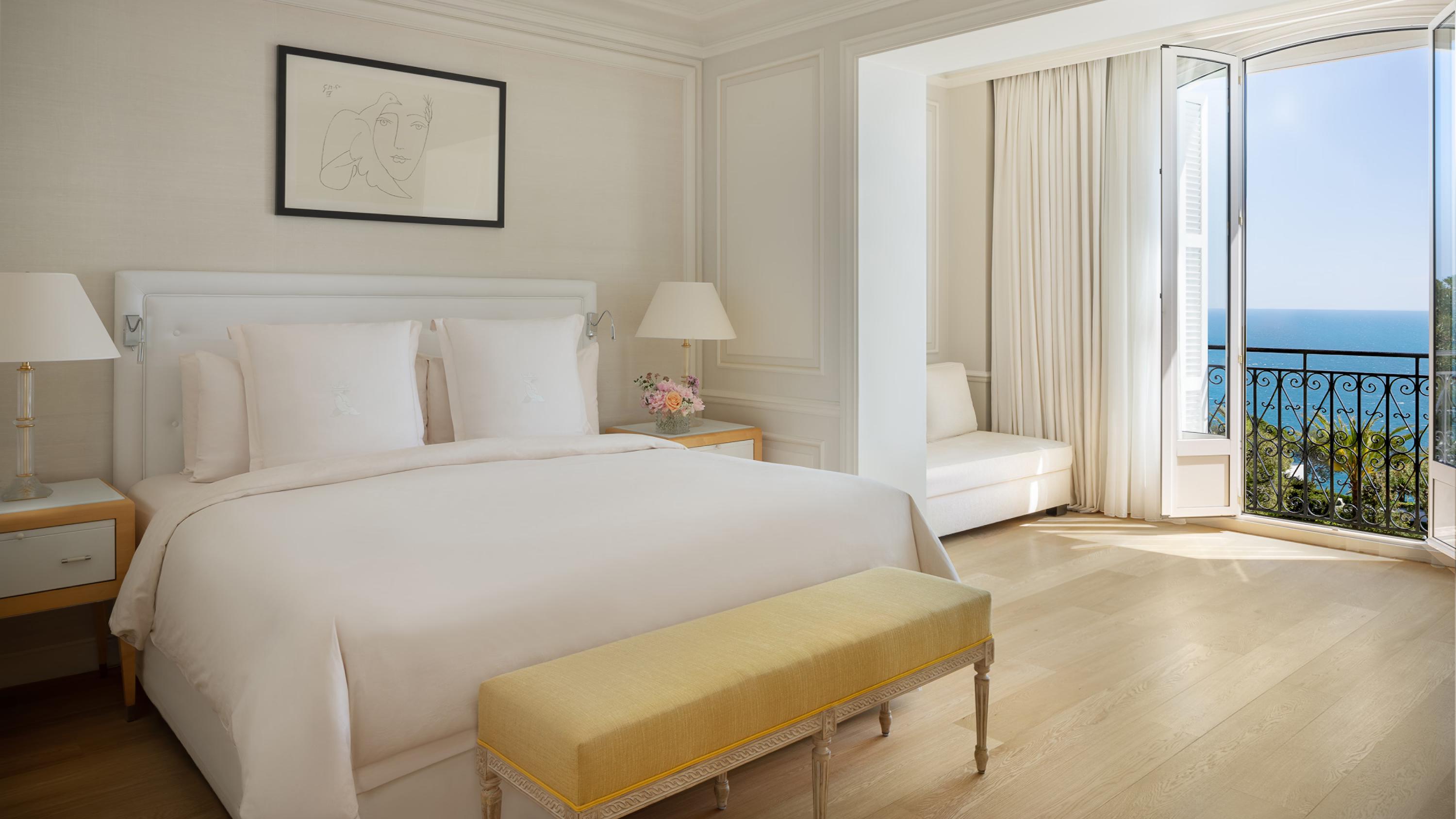 A guest bedroom with a white bed and mustard bench, looking toward a sea-view balcony