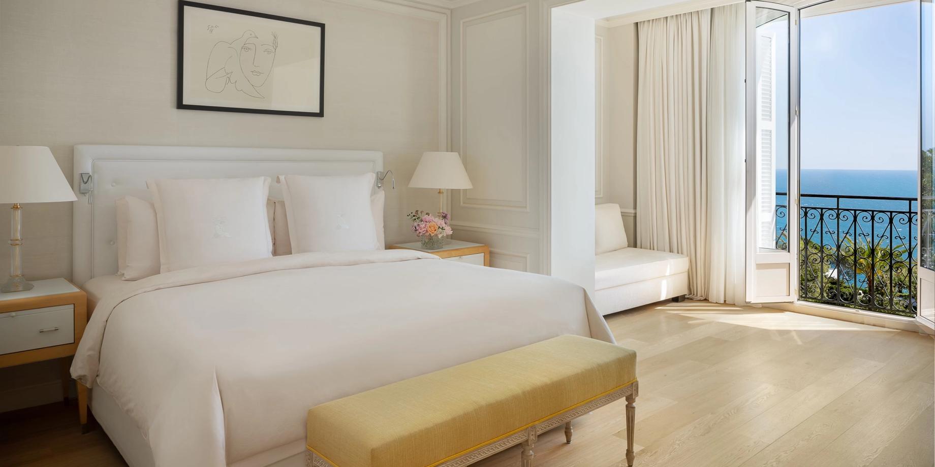 A guest bedroom with a white bed and mustard bench, looking toward a sea-view balcony