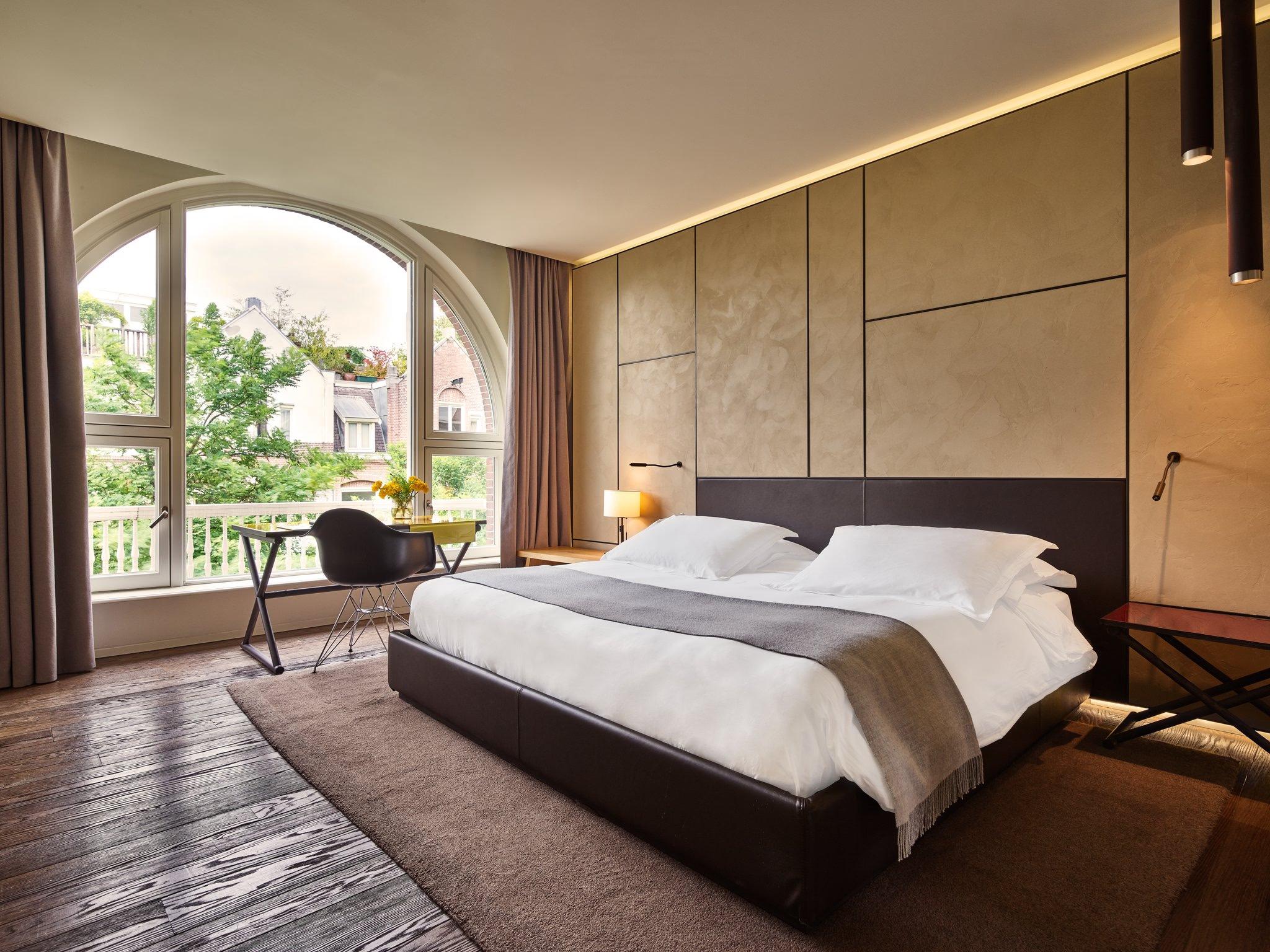 Mandarin Oriental Conservatorium, Amsterdam: bedroom with a tall arched window over the treetops, a desk, and a large bed with a grey throw