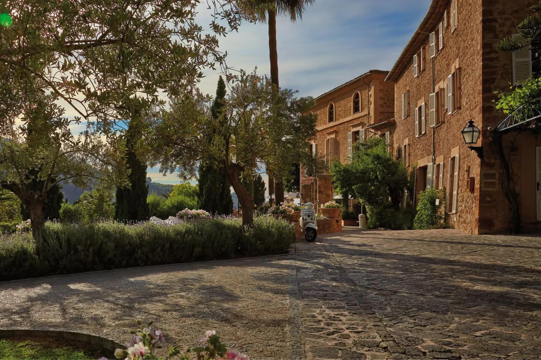 La Residencia: stone pathway between rustic buildings lined with cypress trees and flowering plants