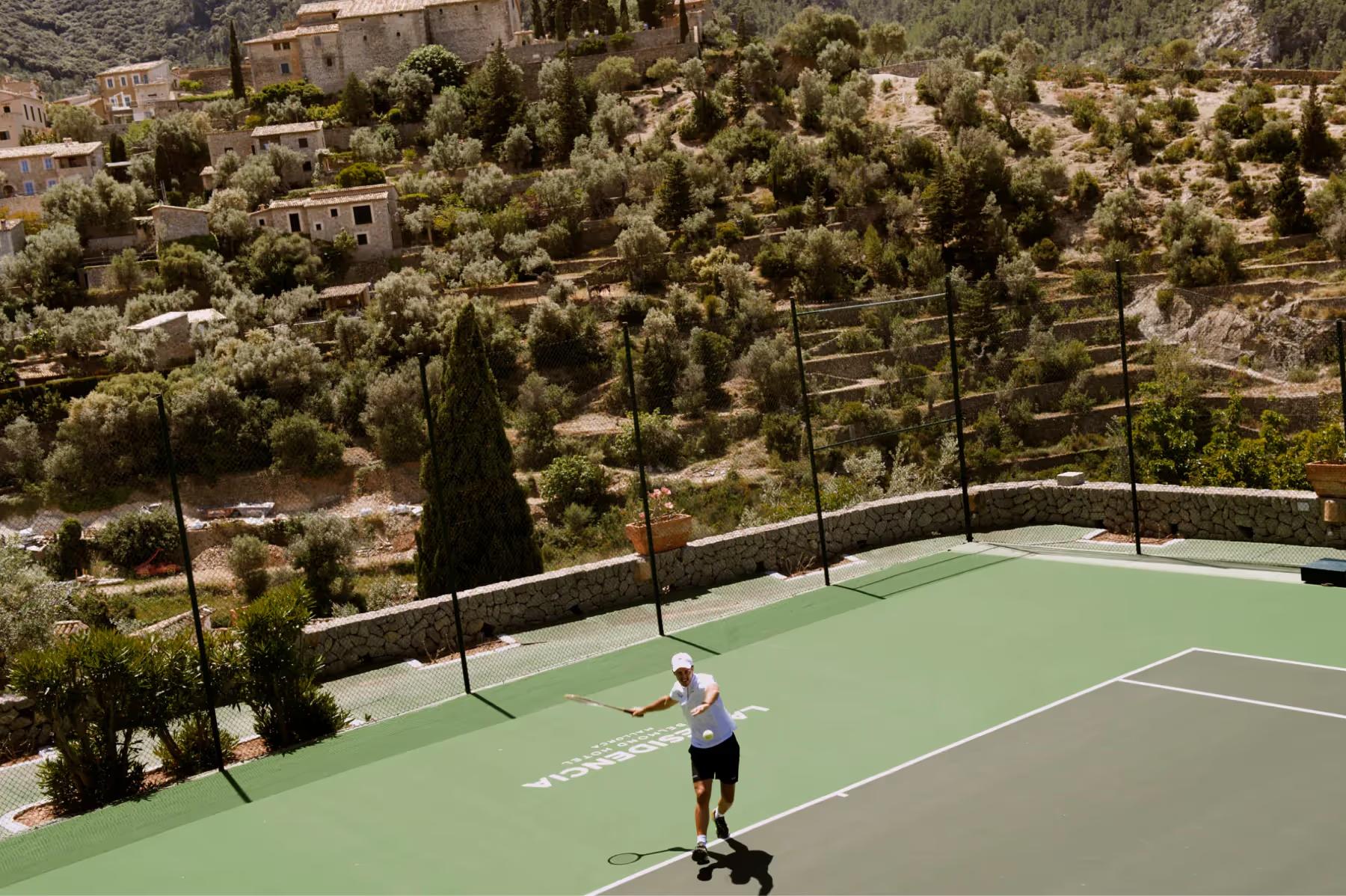 Tennis court on hillside with player, surrounded by Mediterranean vegetation and cypress trees