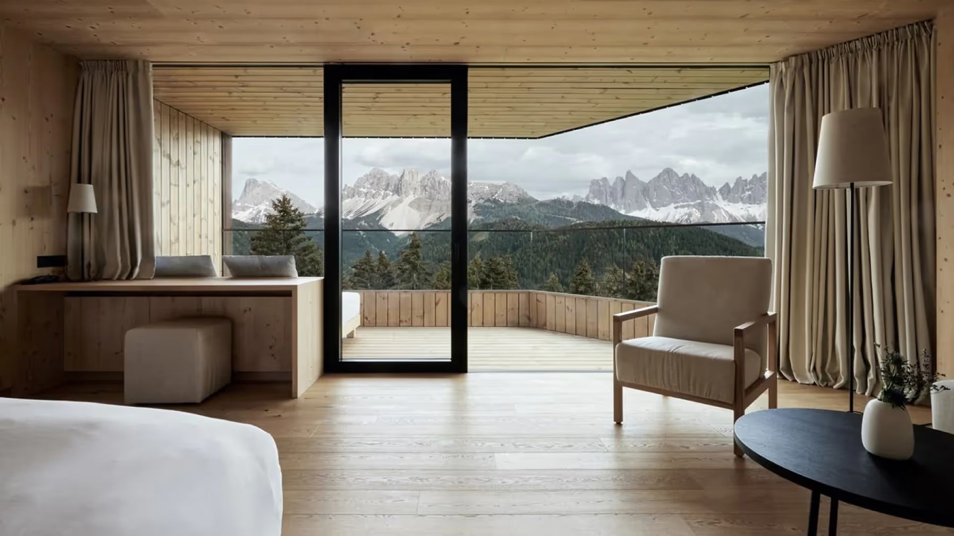 A bright modern suite with expansive windows framing snow-capped mountains; light wood floors, a bed, and stone accents with terrace views