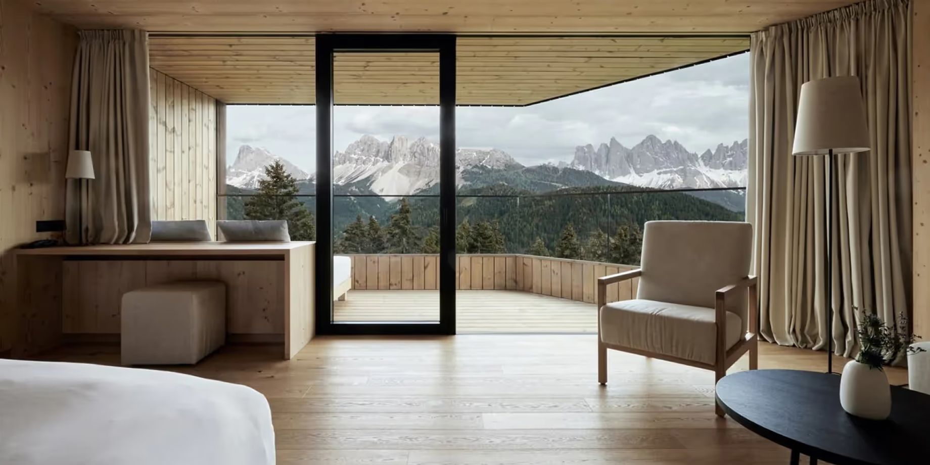 A bright modern suite with expansive windows framing snow-capped mountains; light wood floors, a bed, and stone accents with terrace views