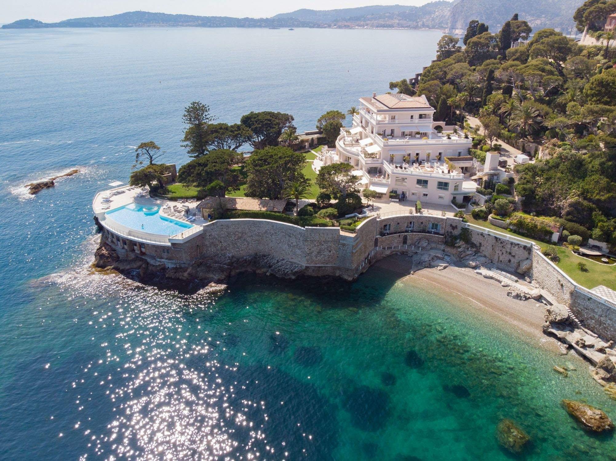 Hotel Cap-Estel: aerial view of a curved white building on a peninsula with turquoise water and sandy beach