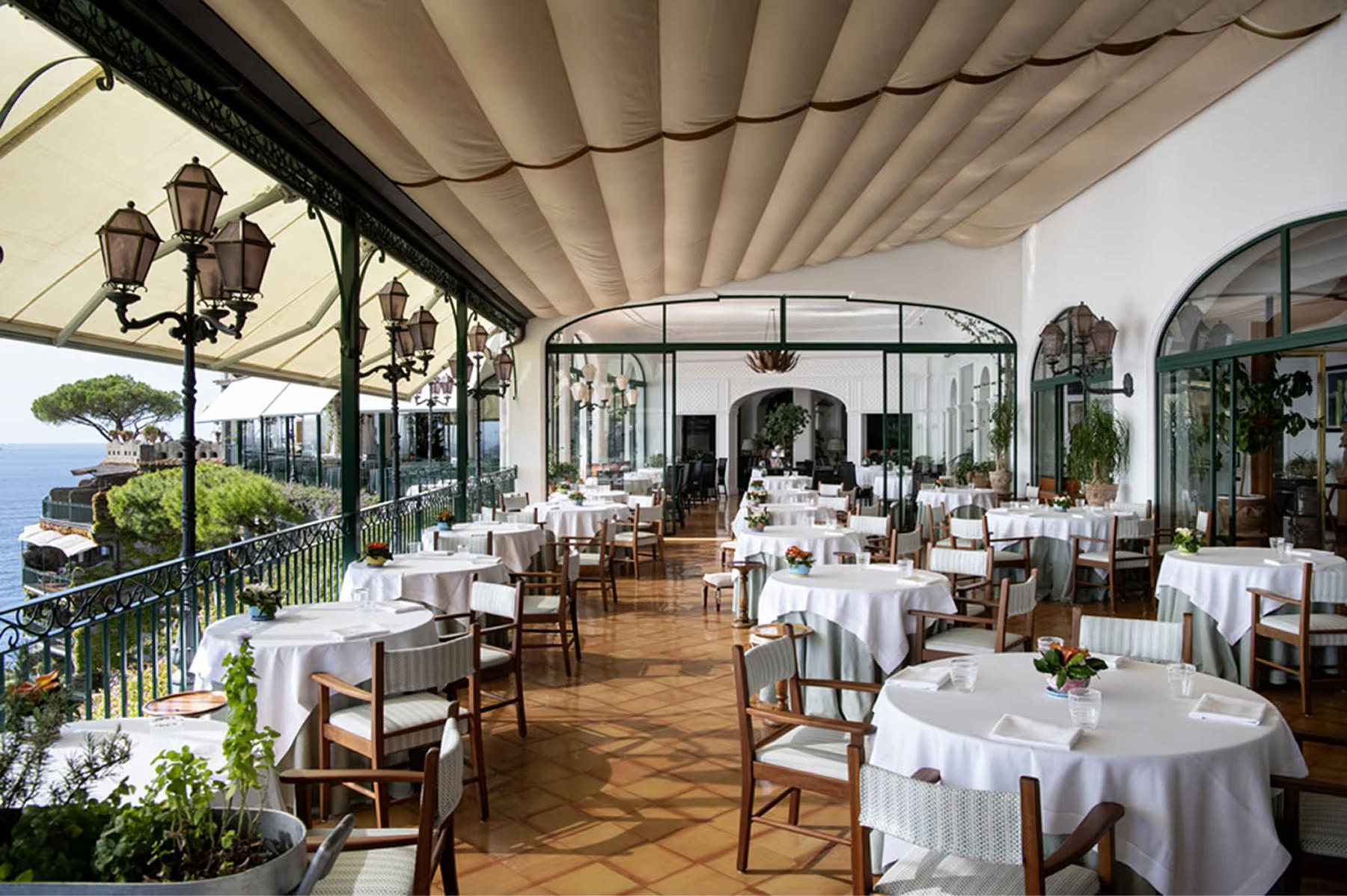 Il San Pietro di Positano: covered terrace dining area with round white-clothed tables, wooden chairs, sea views, Italian coastal setting