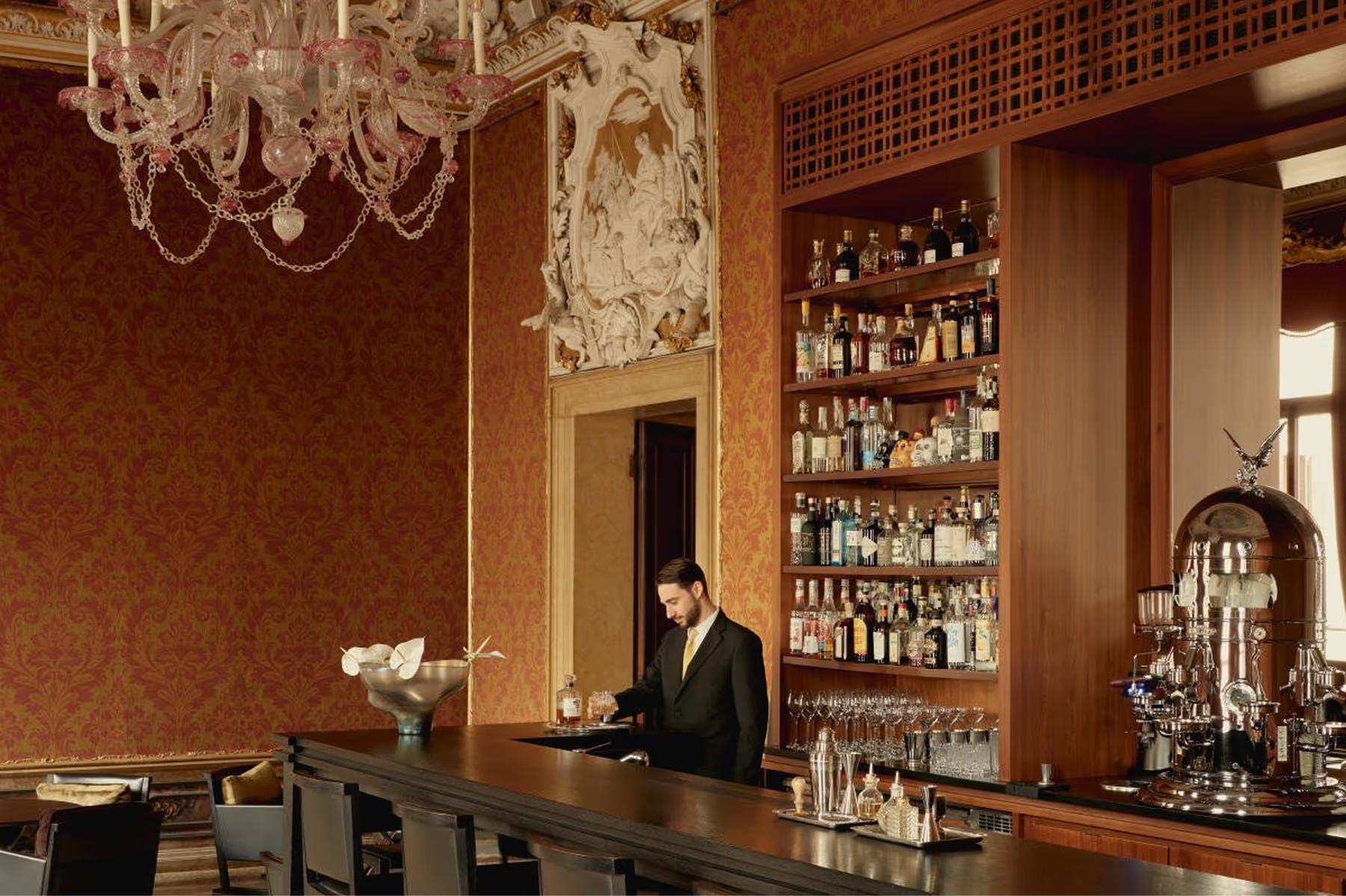 Aman Venice: a classic bar with dark red walls, brass fixtures, a bartender, bottle-lined shelves, and crystal chandelier above
