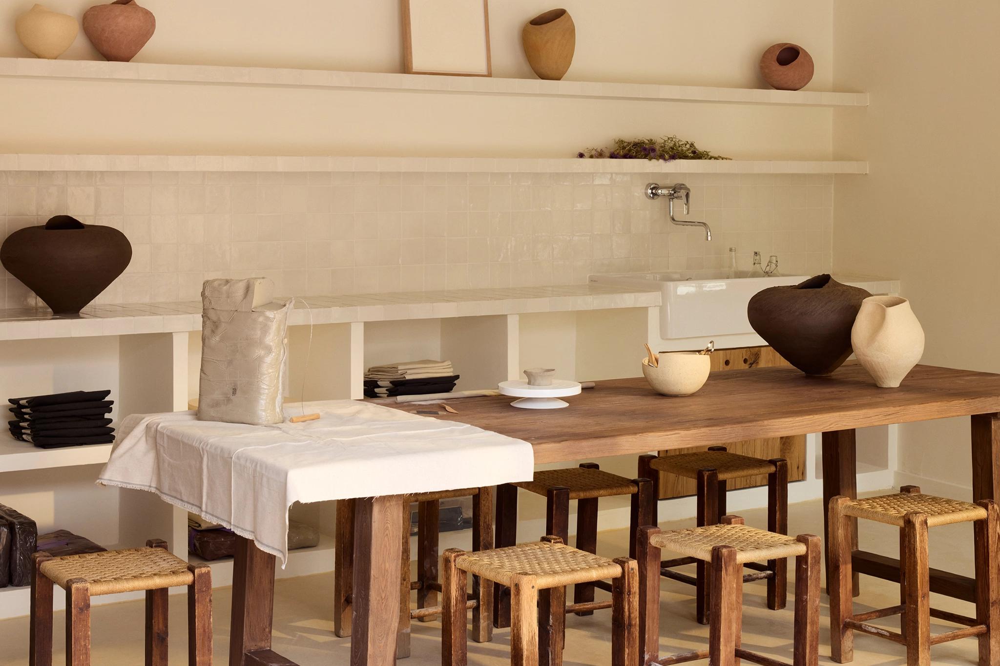 Dark and pale ceramic vessels line a wooden table beneath a tiled backsplash and shelf