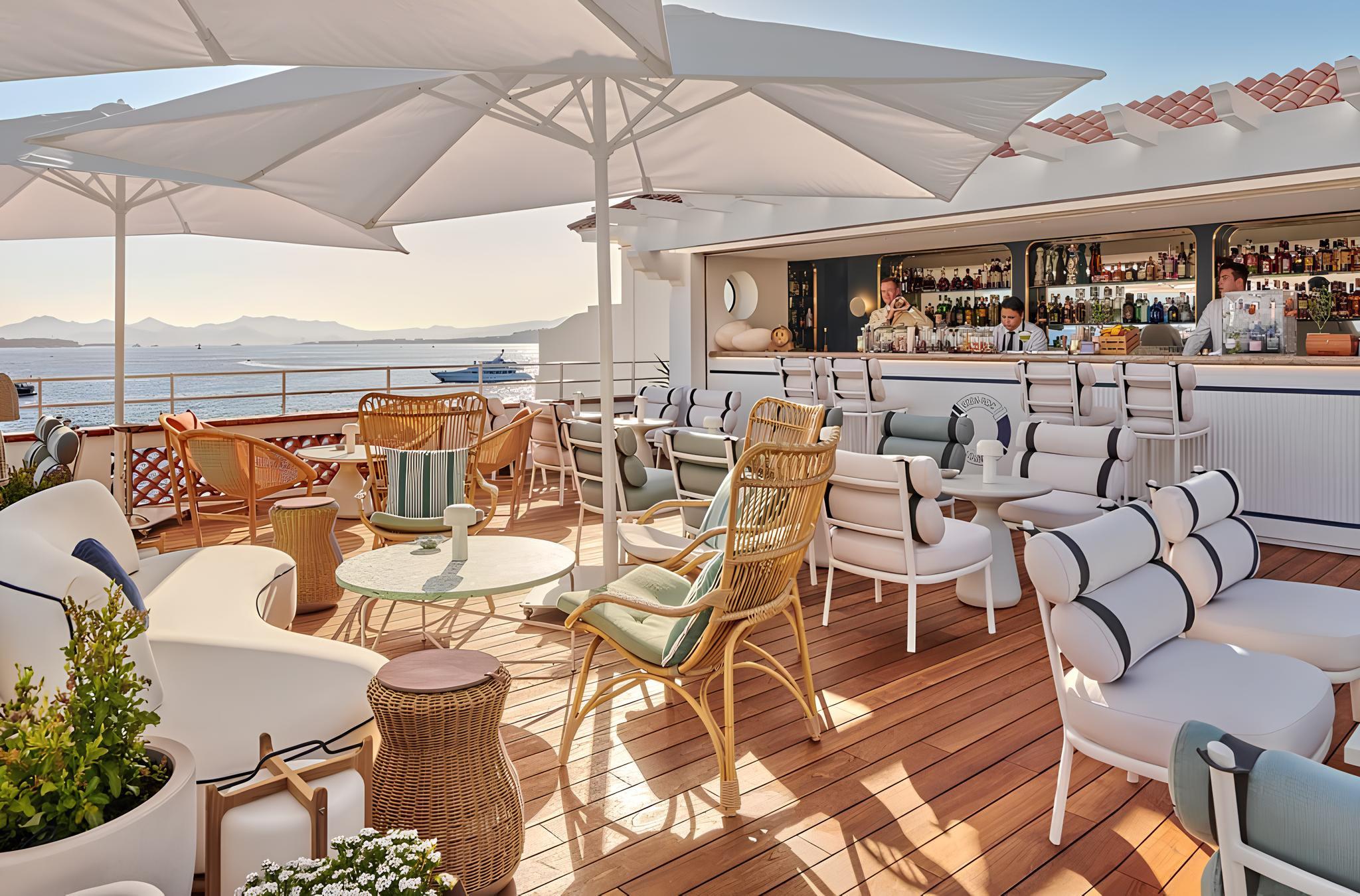 A sun-drenched terrace with white umbrellas, wooden deck, bar area, and Mediterranean panorama