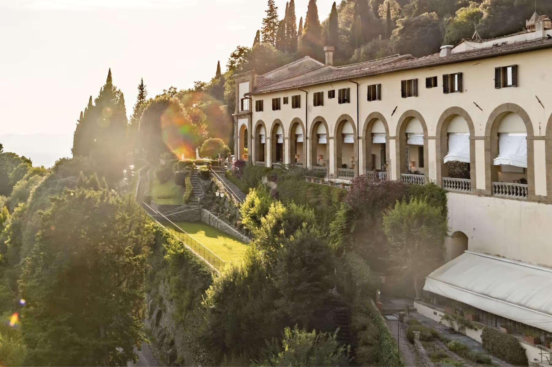 Cream building with arched columns, shuttered windows, cypress trees, and golden light across the hillside