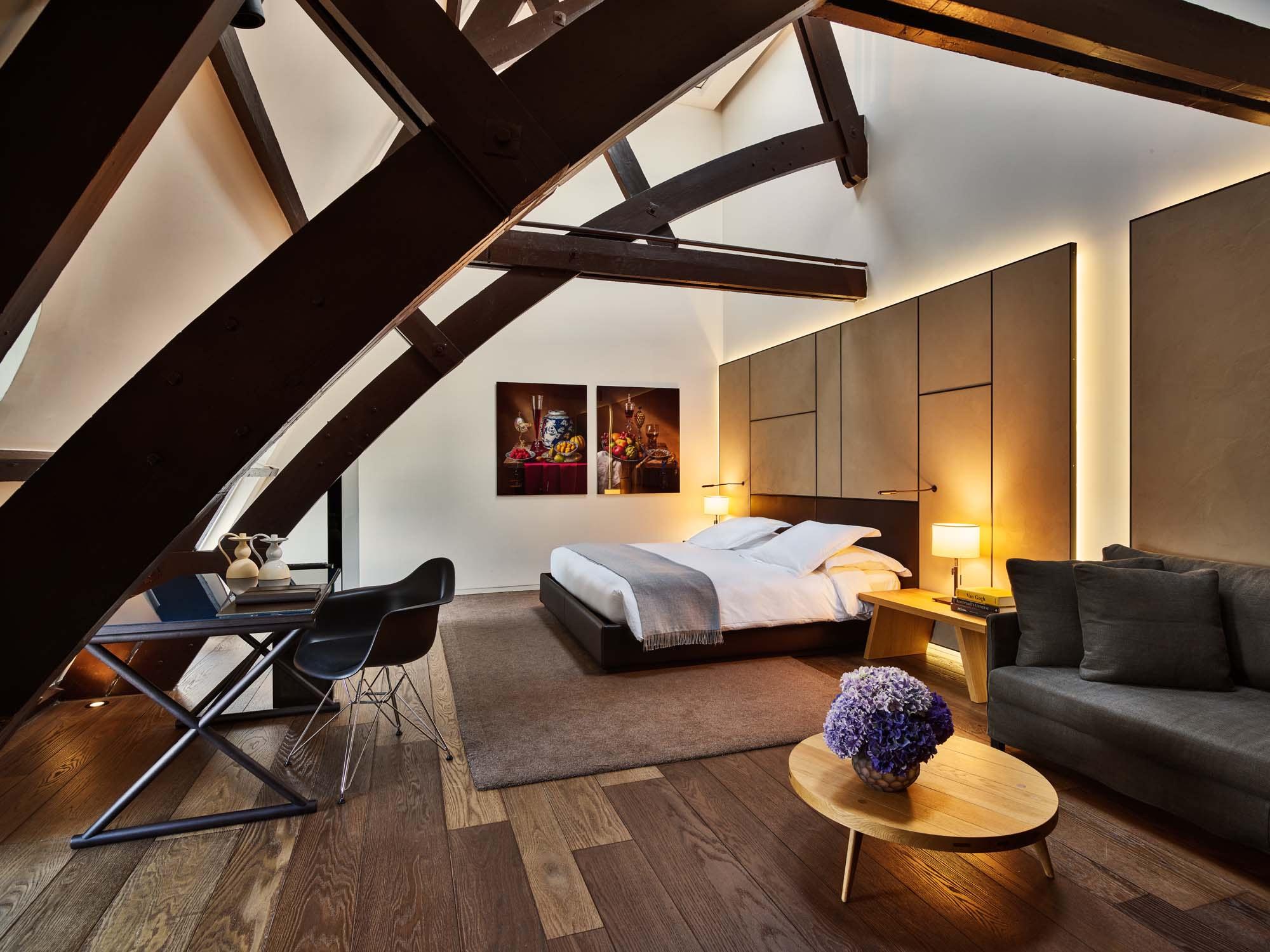 Mandarin Oriental Conservatorium, Amsterdam: bedroom under exposed timber roof beams, with a desk, a grey sofa, and an oval table with purple flowers