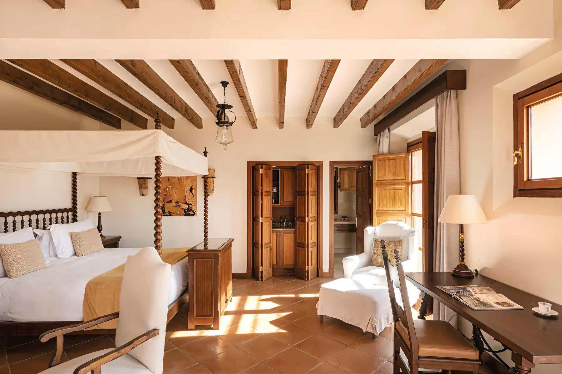 Sun-filled hotel bedroom with exposed beam ceiling, ornate wooden furnishings, and archway entrance