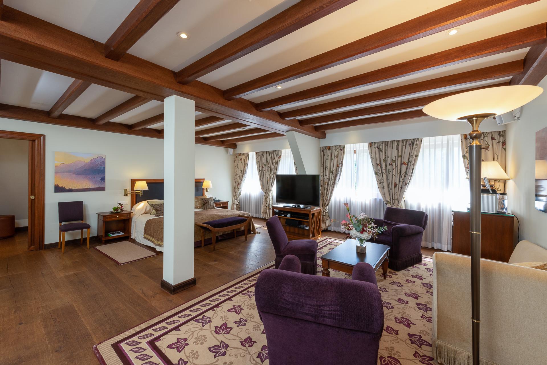 A junior suite with exposed wooden beams, purple armchairs, and corner windows overlooking the valley