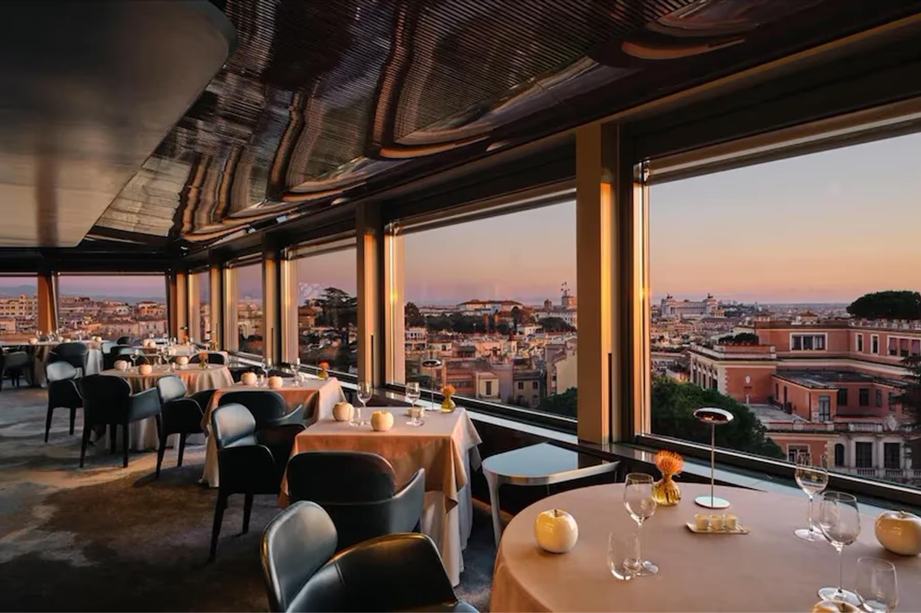 Hotel Eden: La Terrazza rooftop restaurant at dusk with floor-to-ceiling windows, diners at round tables, Rome’s skyline in the background