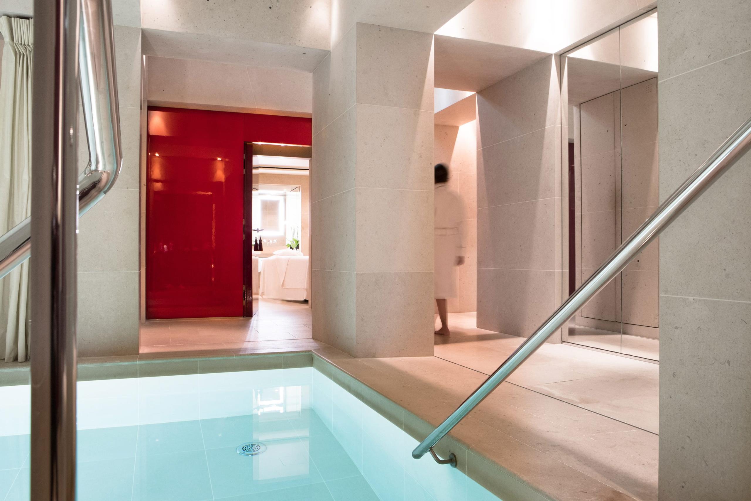 A spa corridor with glass partitions, red walls, and a soaking tub visible through doorway