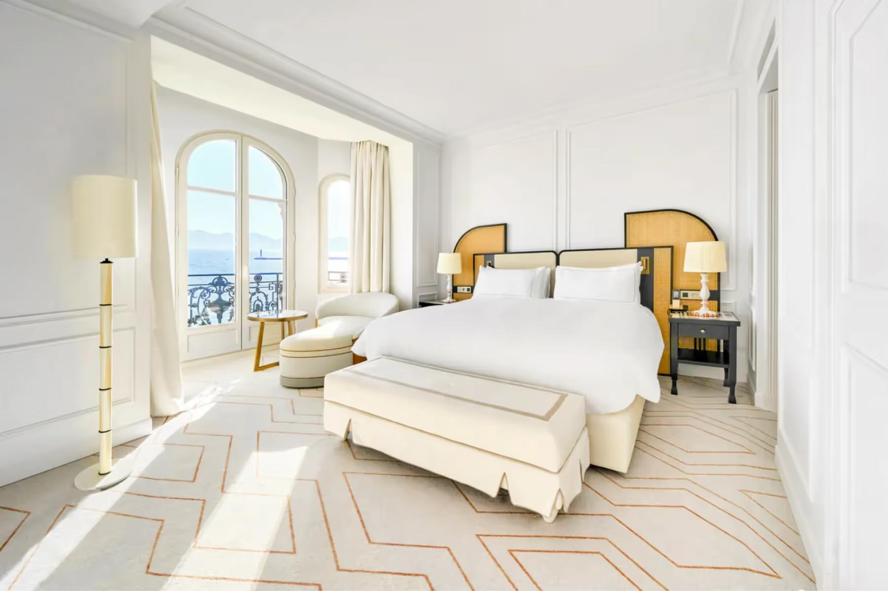 Carlton Cannes: bedroom with white bed, brass-trimmed nightstands, geometric floor pattern, and glass doors to a balcony with sea views