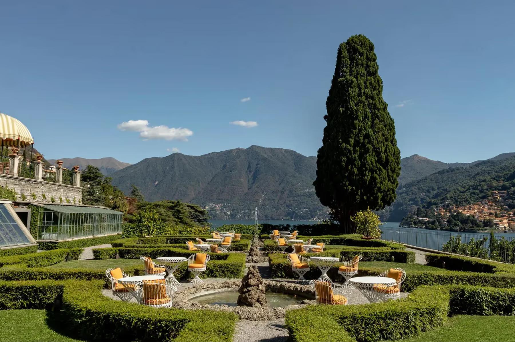 Manicured grounds with a tall cypress, scattered seating, and lake views beyond hedges