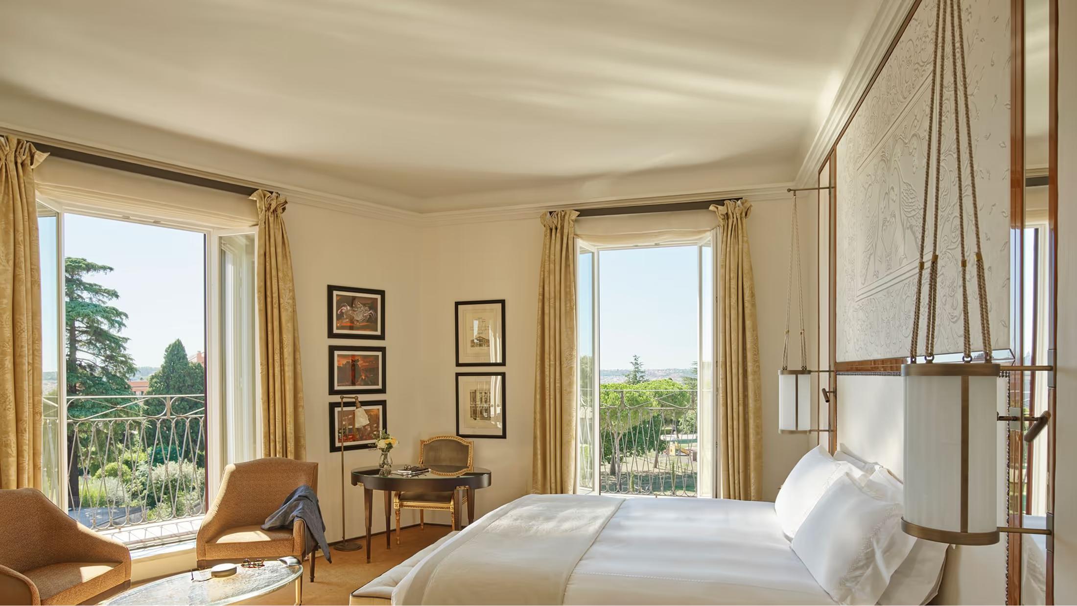 A bedroom with pale stone walls, tall windows overlooking Rome, and natural light illuminating the bed
